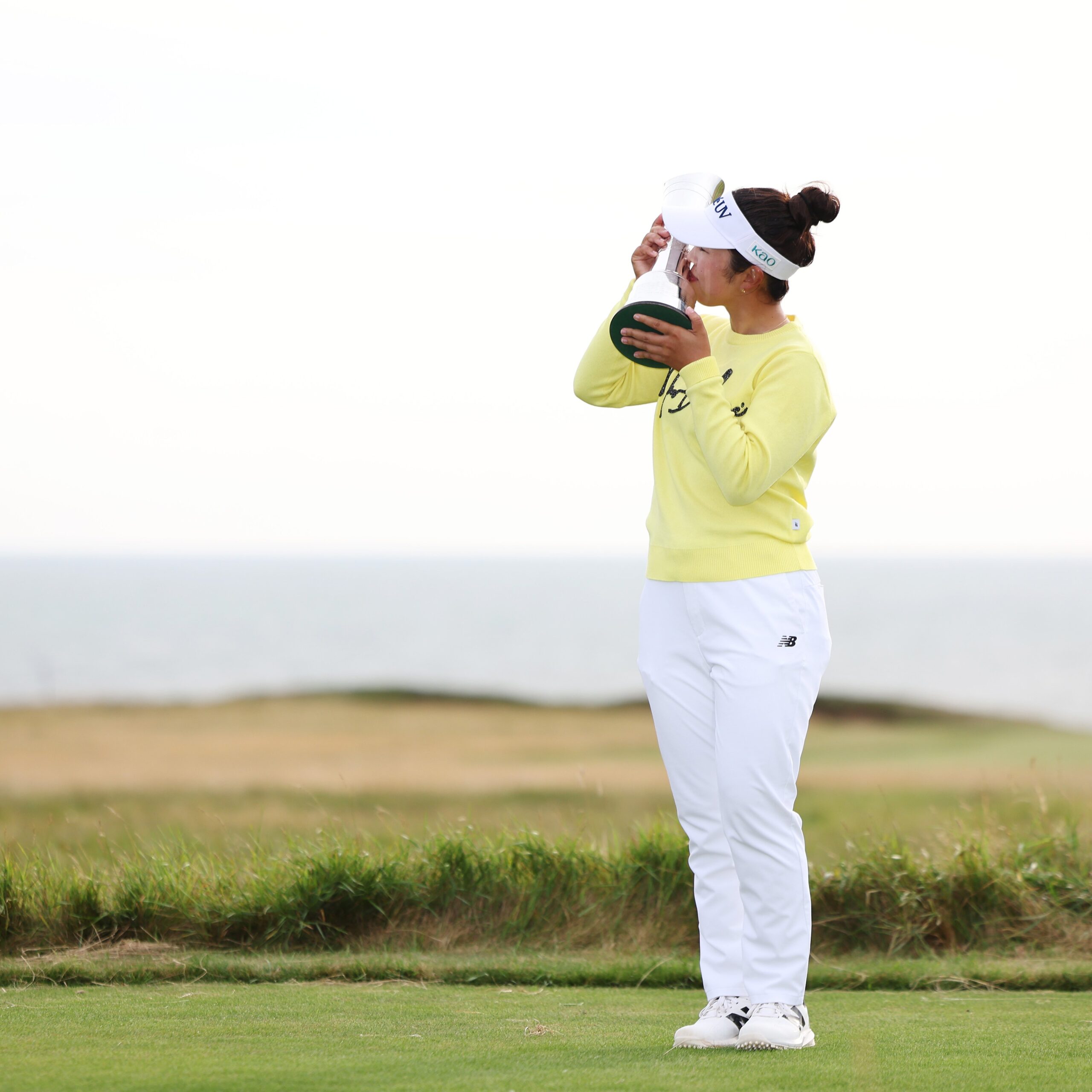 Miyu Yamashita, wearing a butter yellow-green top and white pants, holds the AIG Women's Open trophy up to her right and kisses the side of it. The backdrop of Royal Porthcawl in Wales is in the distance.