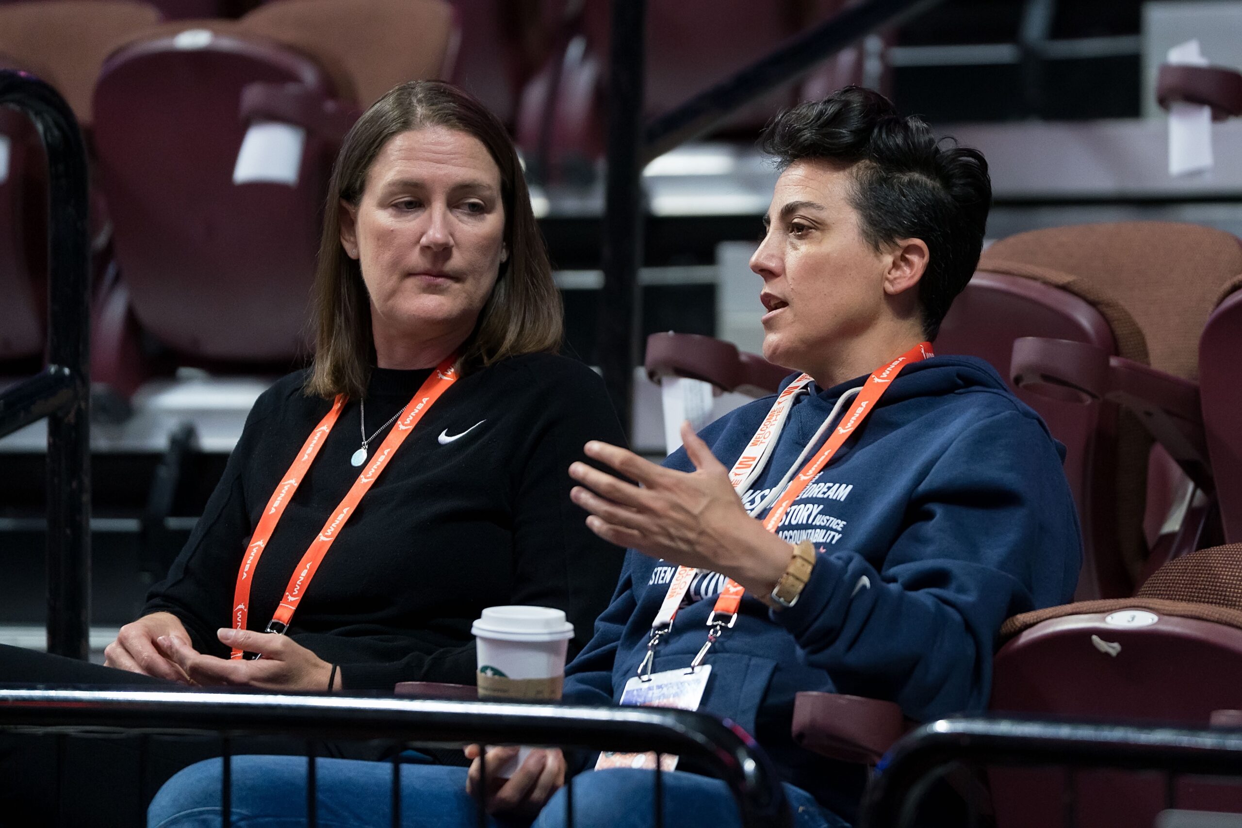 Washington Mystics general manager Jamila Wideman and assistant general manager Maria Giovannetti sit in the stands and talk before a game. Wideman holds a coffee cup in her right hand and gestures with her left as she speaks.