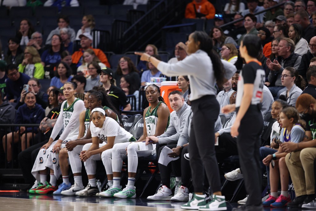 Seattle Storm head coach Noelle Quinn is blurry in the foreground pointing to something on the court. On the bench, Zia Cooke and Nneka Ogwumike look toward her and the rest of the players look toward the court.