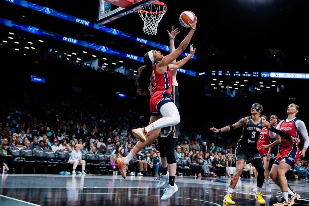 Washington Mystics forward Kiki Iriafen is shown in midair. She is on the left side of the basket with her back to the baseline, and she extends her right arm to try to flip the ball behind her for a reverse layup.
