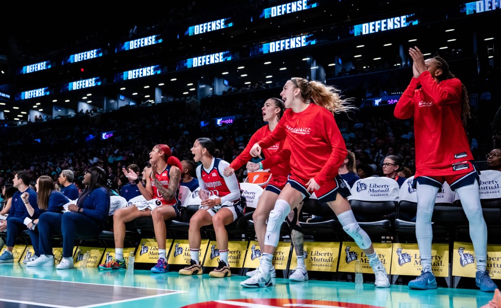 Washington Mystics players celebrate on the bench. Two yell and clap while seated, and three others are on their feet cheering.