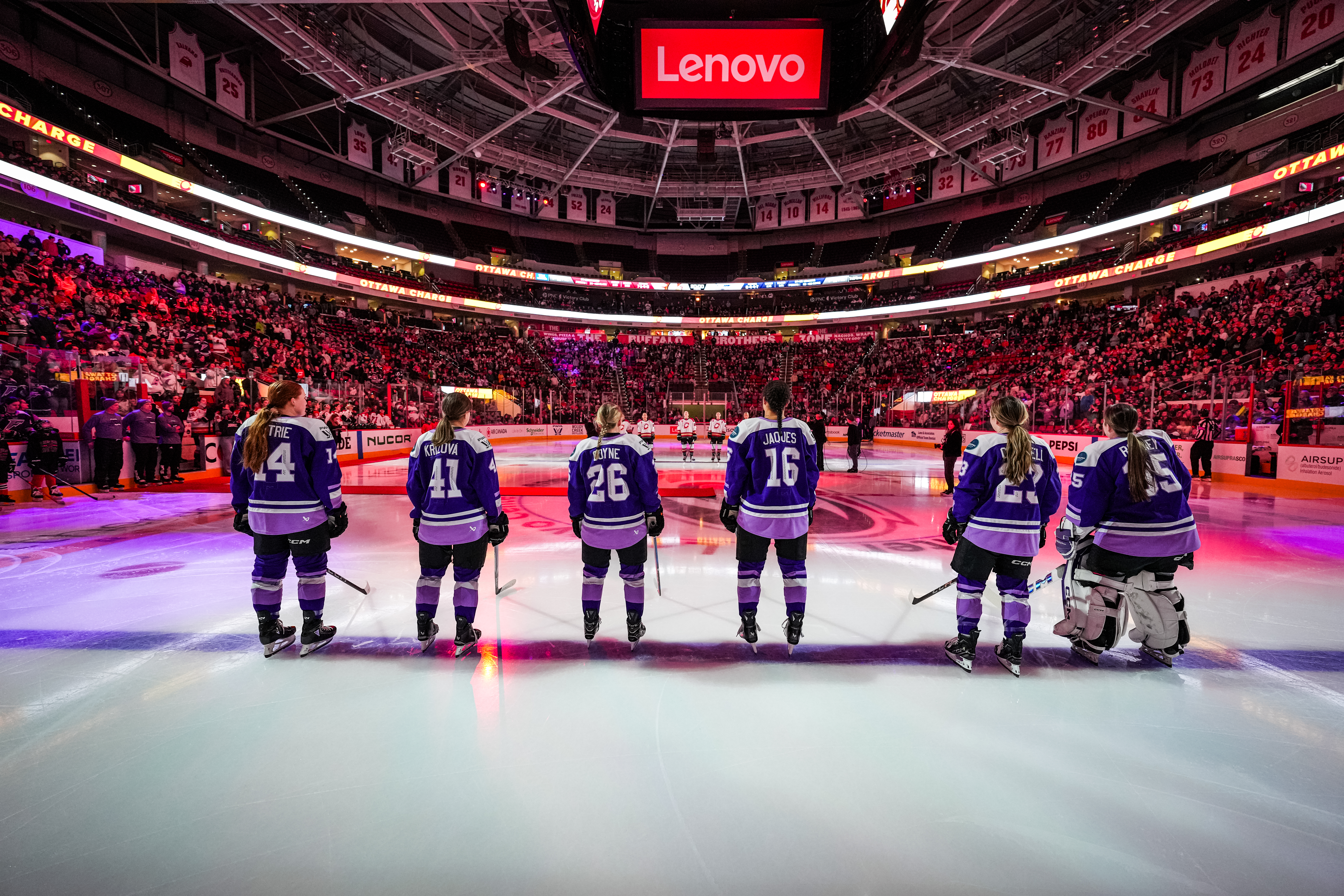Five skaters and a goaltender stand across the blueline with their backs toward the camera. They are all wearing purple home uniforms.