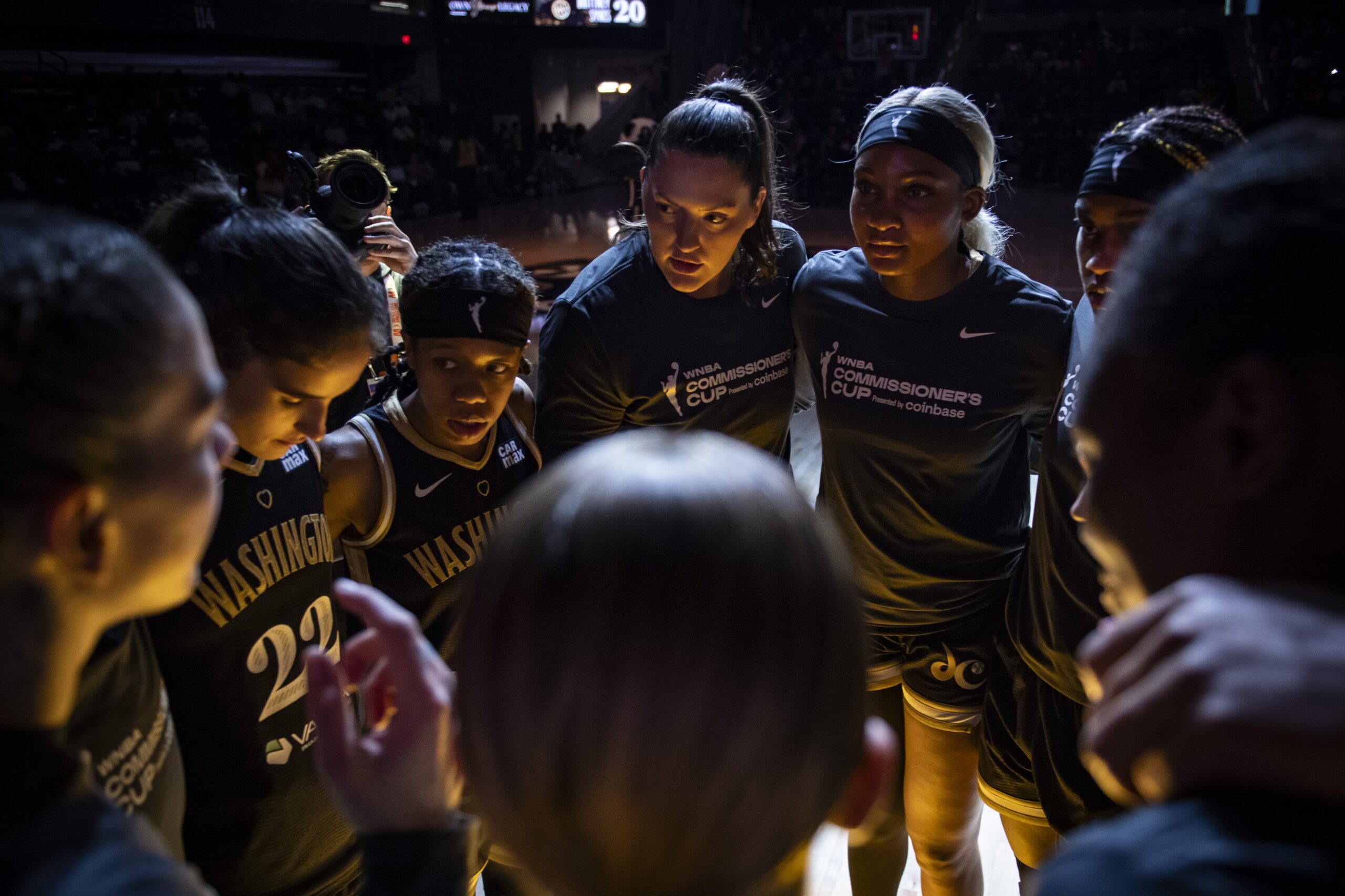 The lights are dark at CareFirst Arena as Washington Mystics players huddle before a game. They are wearing black jerseys with gold trim or black warm-up shirts.