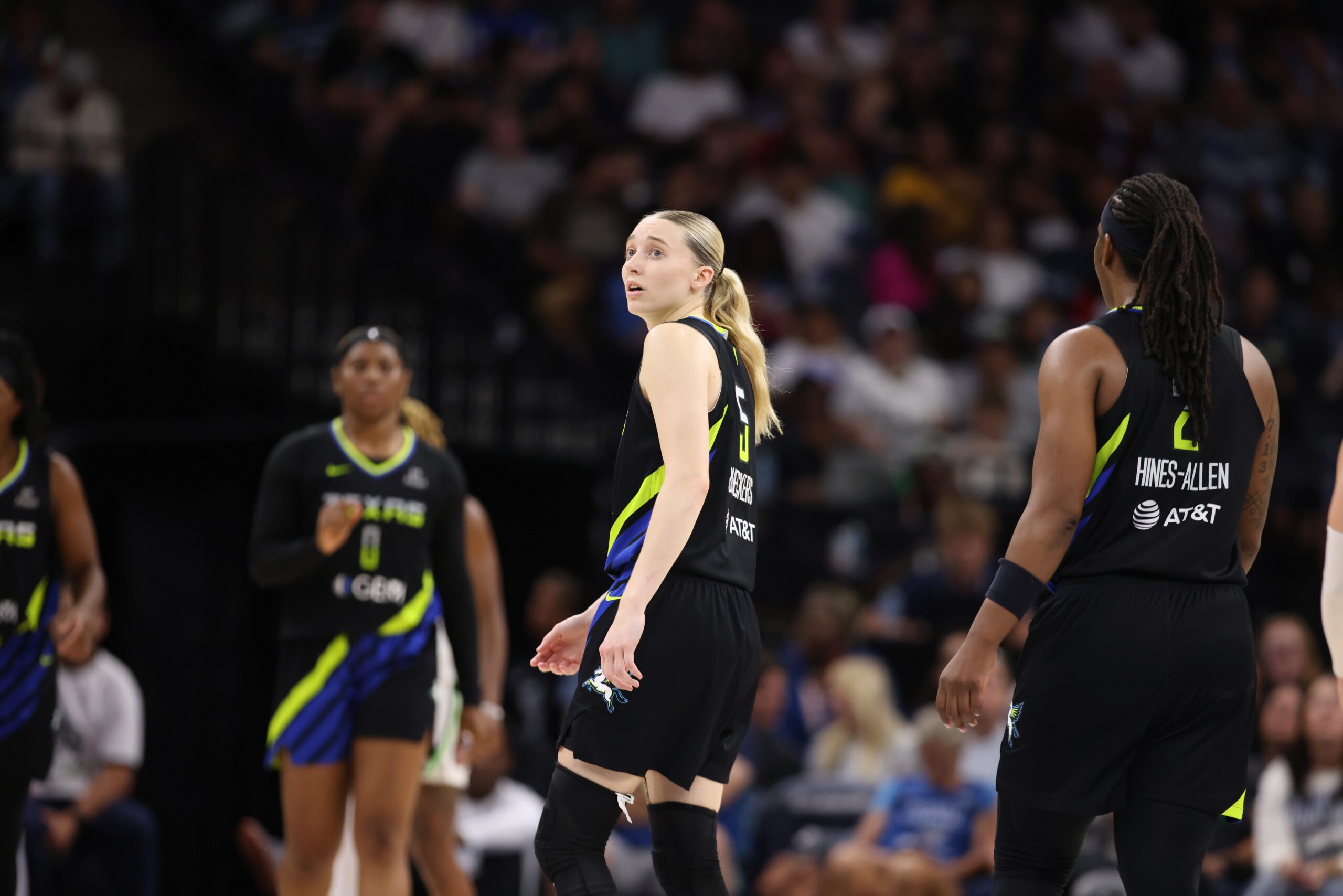 Dallas Wings guard Paige Bueckers looks up with a concerned expression. Teammate Myisha Hines-Allen approaches her from the side, while other teammates look on in the background. Packed arena stands are blurry in the background.