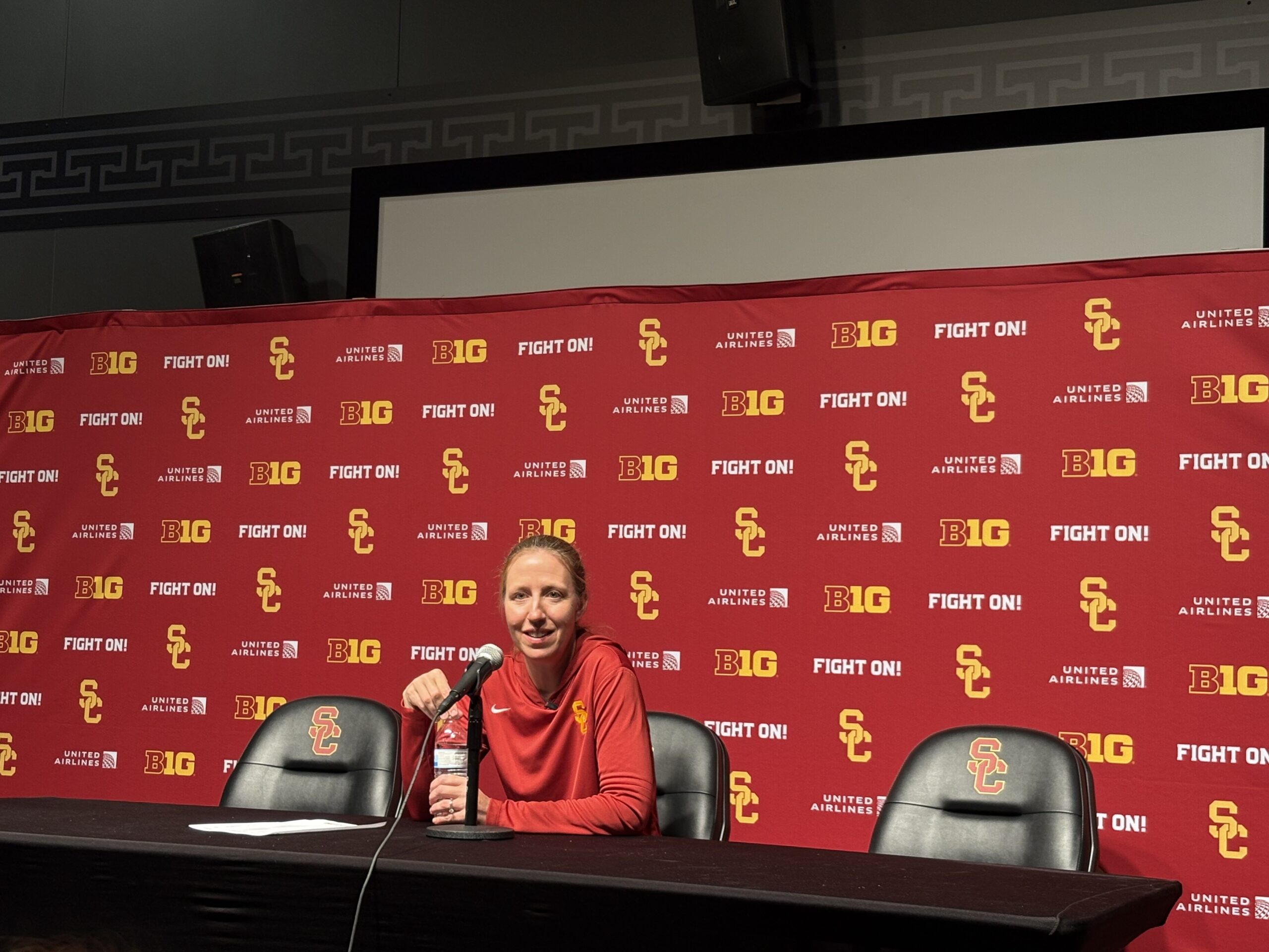 USC Head Coach Lindsay Gottlieb speaks to the media in front of a red banner reading "Fight On!" and SC, smiling as she answers questions.
