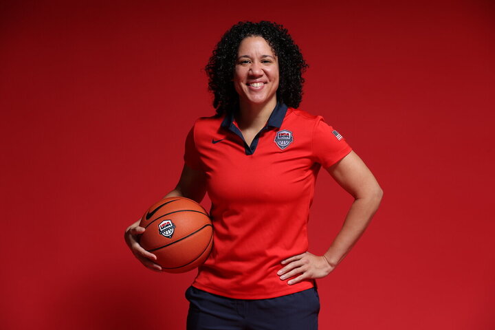 Kara Lawson stands with a ball tucked under arm in a red USA Basketball polo