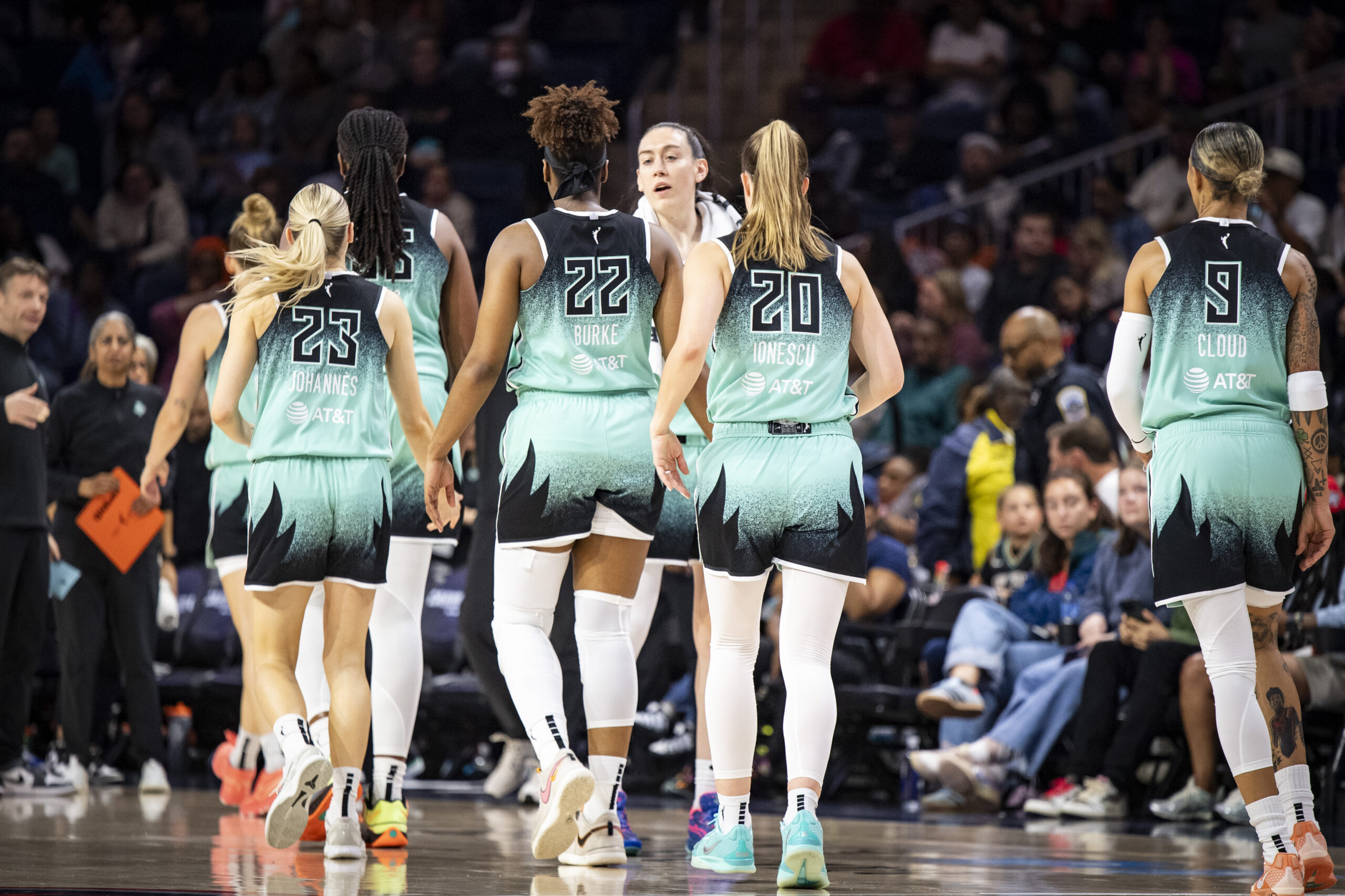 Members of the New York Liberty walk away down the court, including Marine Johannès, Jonquel Jones, Kennedy Burke, Sabrina Ionescu, and Natasha Cloud, toward Breanna Stewart who has a towel around her neck. The Liberty coaching staff waits in the background, in front of an out-of-focus packed crowd.