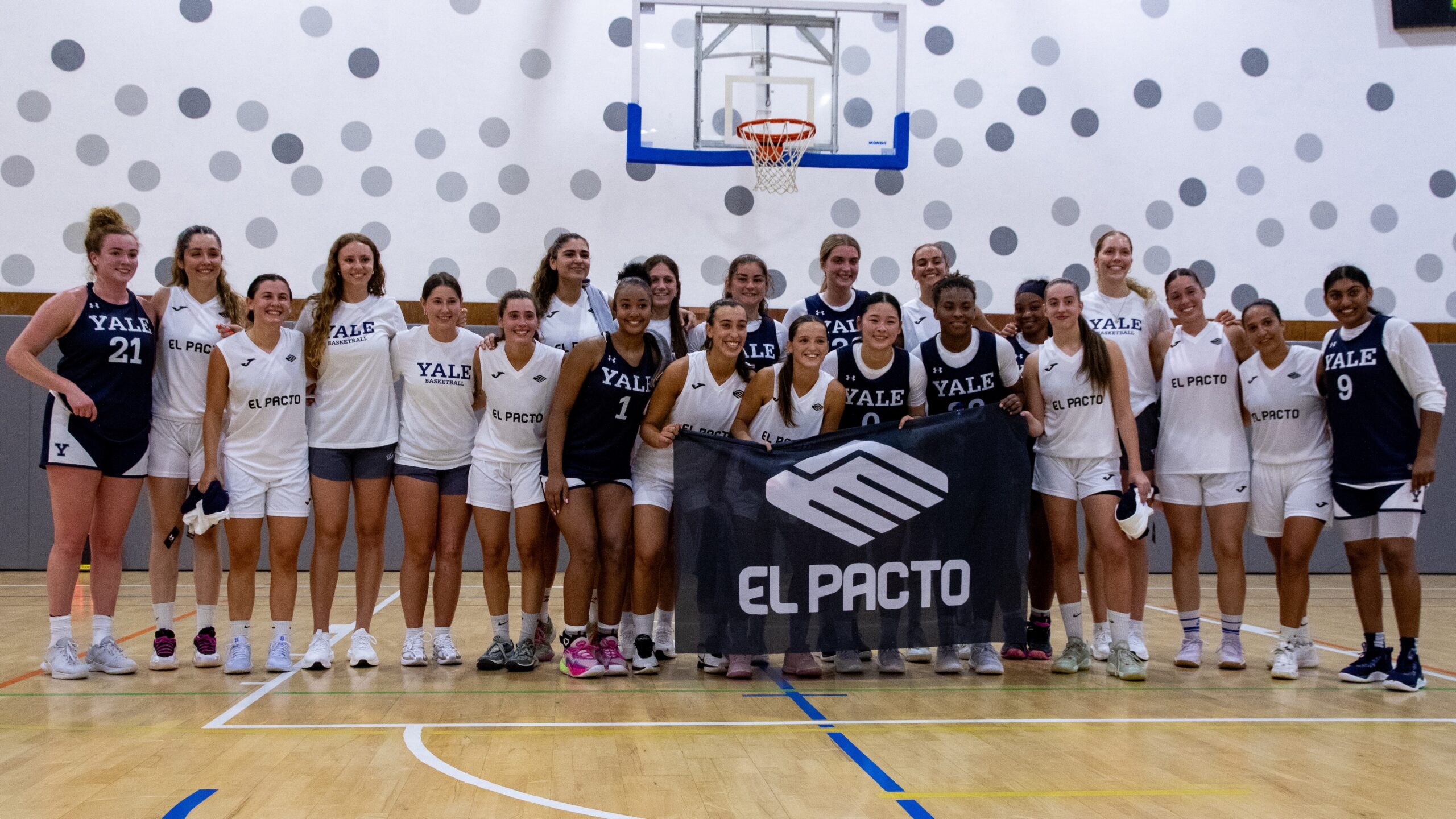 Yale and El Pacto women's basketball players pose for a photo. They are interspersed in two rows, and the players in the front row are holding a black flag that says "El Pacto" on it in white capital letters.