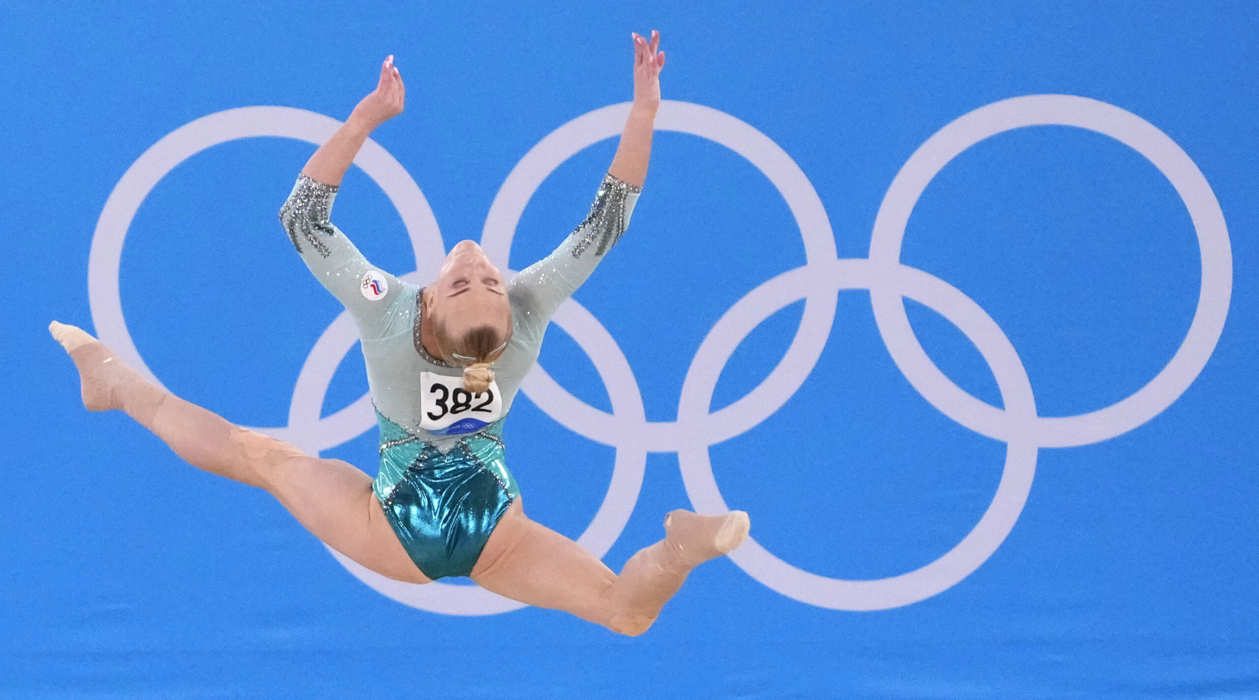 A gymnast in a teal leotard leaps in front of the Olympic rings.
