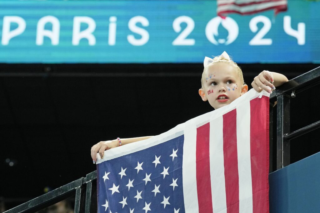 A young fan drapes an American flag over a railing while watching something out of frame, with the "Paris 2024" Olympic wordmarks on a banner in the background