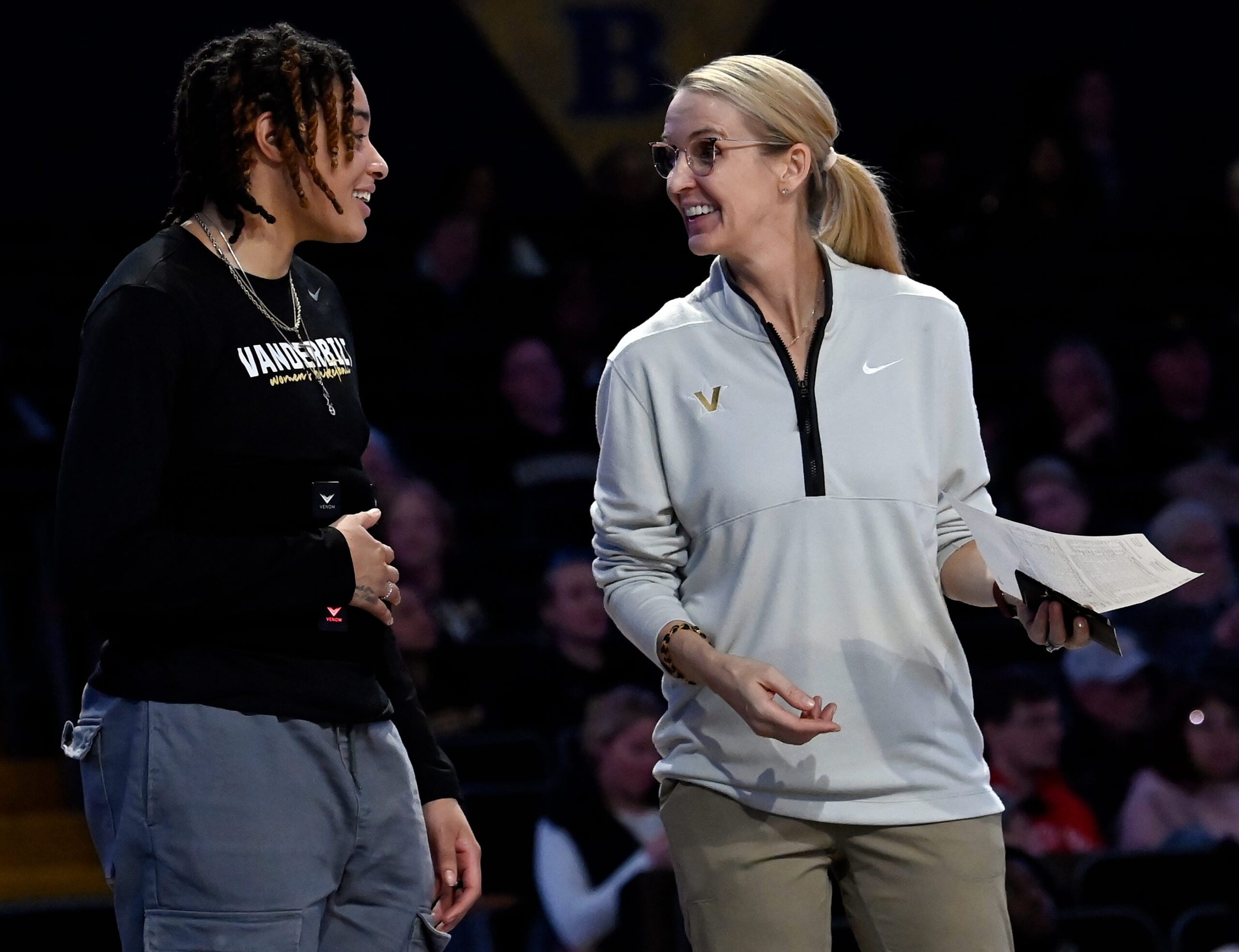 A player on the left wearing black talks with her coach on the right wearing a gray quarter zip. They are smiling in conversation with one another.