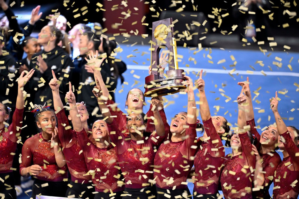 A gymnastics team dressed in red leotards celebrates a victory as confetti falls round them. 