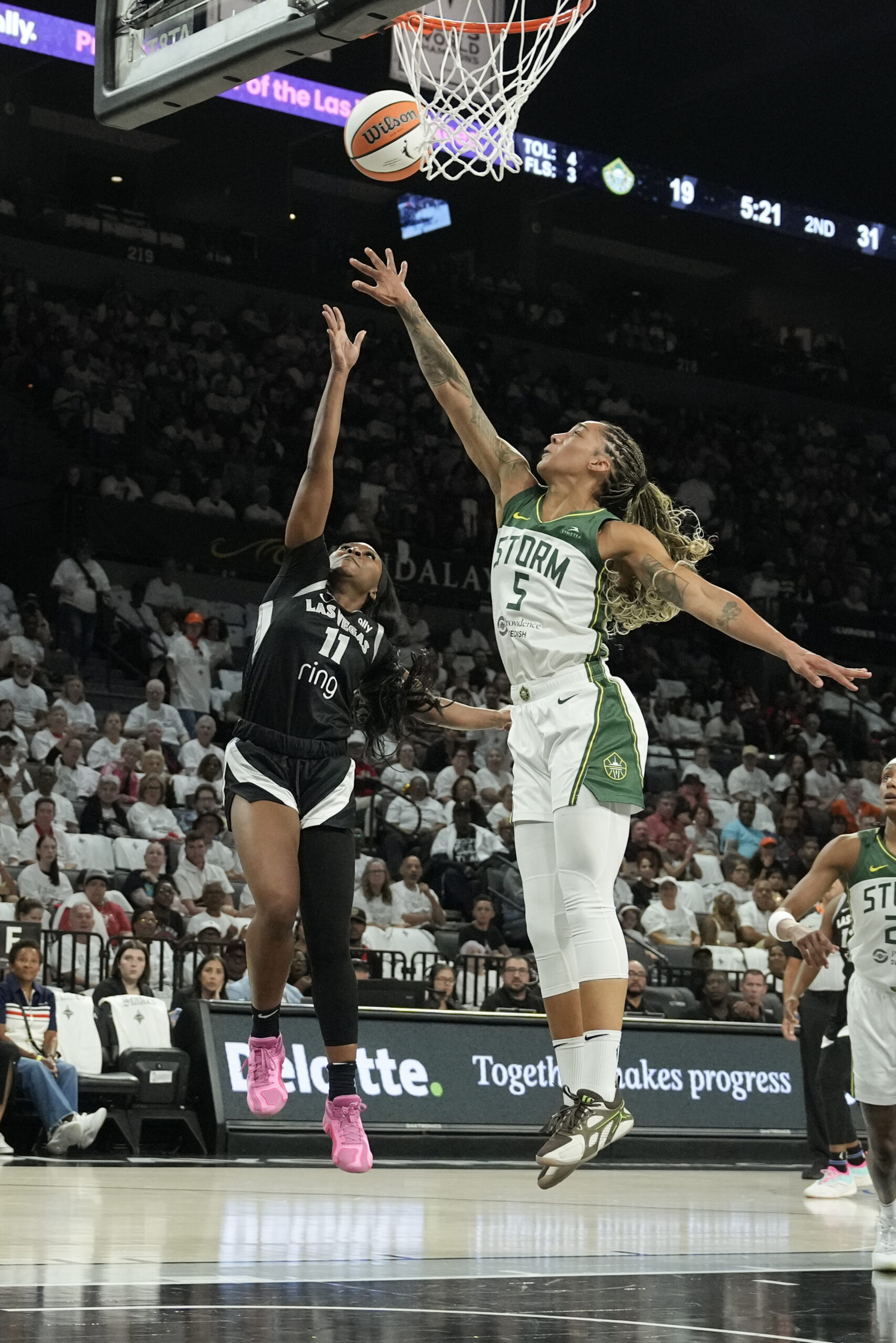 Two players jump for a ball near the basket, one in a black uniform and the other in a white and green uniform.