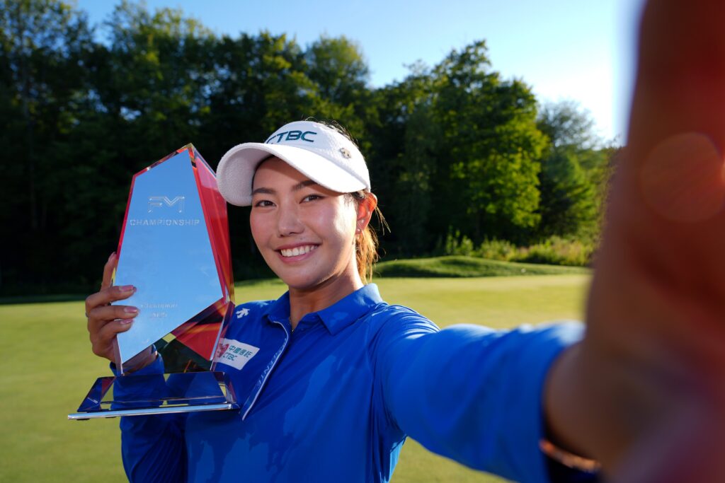 Miranda Wang, winner of the 2025 FM Championship, takes a selfie with the trophy. She holds the camera with her left arm outstretched, and she holds the FM Championship trophy up with her right hand.