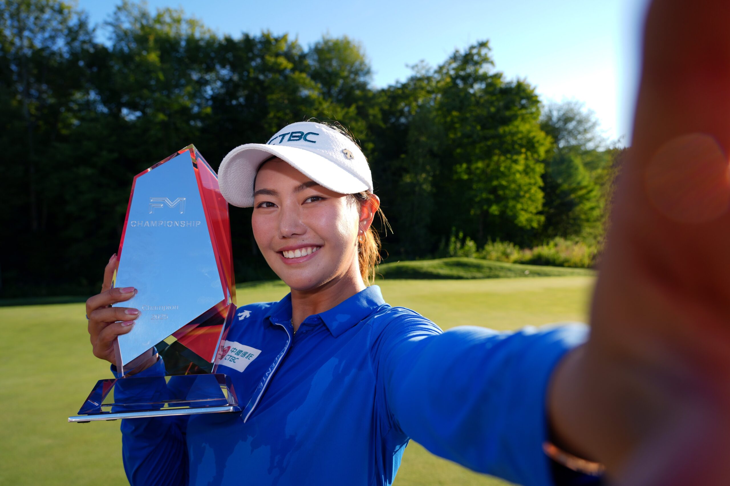 Miranda Wang, winner of the 2025 FM Championship, takes a selfie with the trophy. She holds the camera with her left arm outstretched, and she holds the FM Championship trophy up with her right hand.
