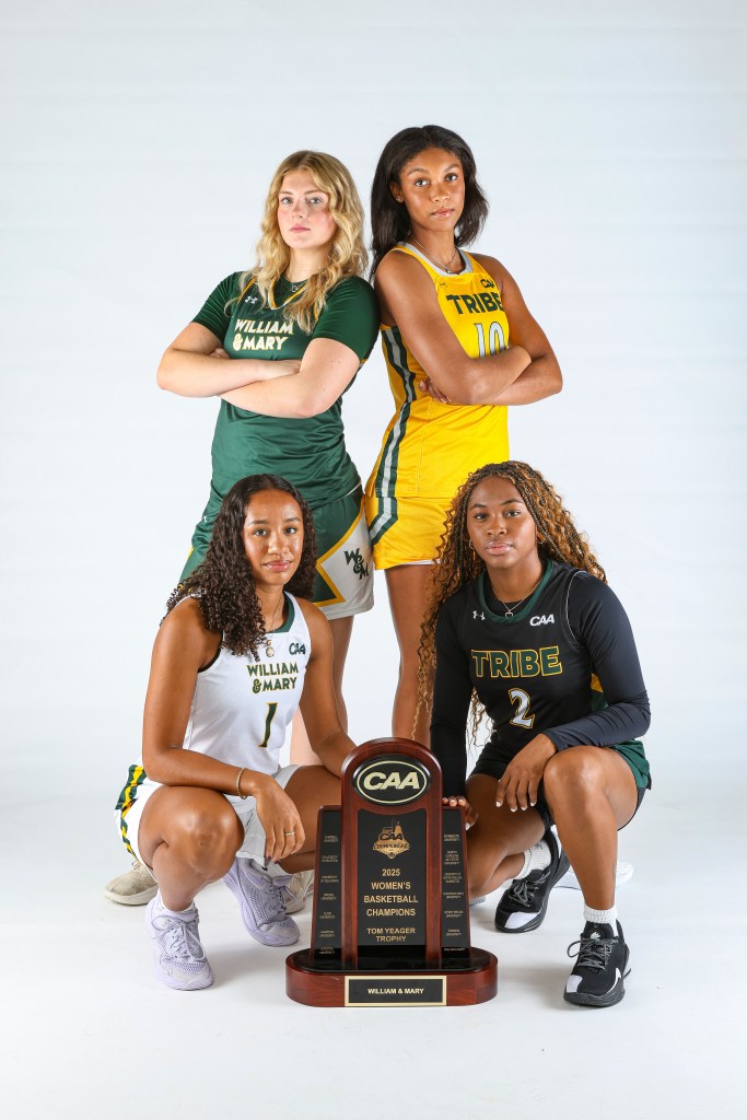 William & Mary women's basketball players Emma West, Marley Long, Cassidy Geddes and Monet Dance pose for a photo. West, wearing a green uniform, and King in a gold jersey are standing shoulder to shoulder with their backs to each other. Wearing a white jersey, Geddes and Dance, in a black uniform, are kneeling down with the CAA championship trophy.