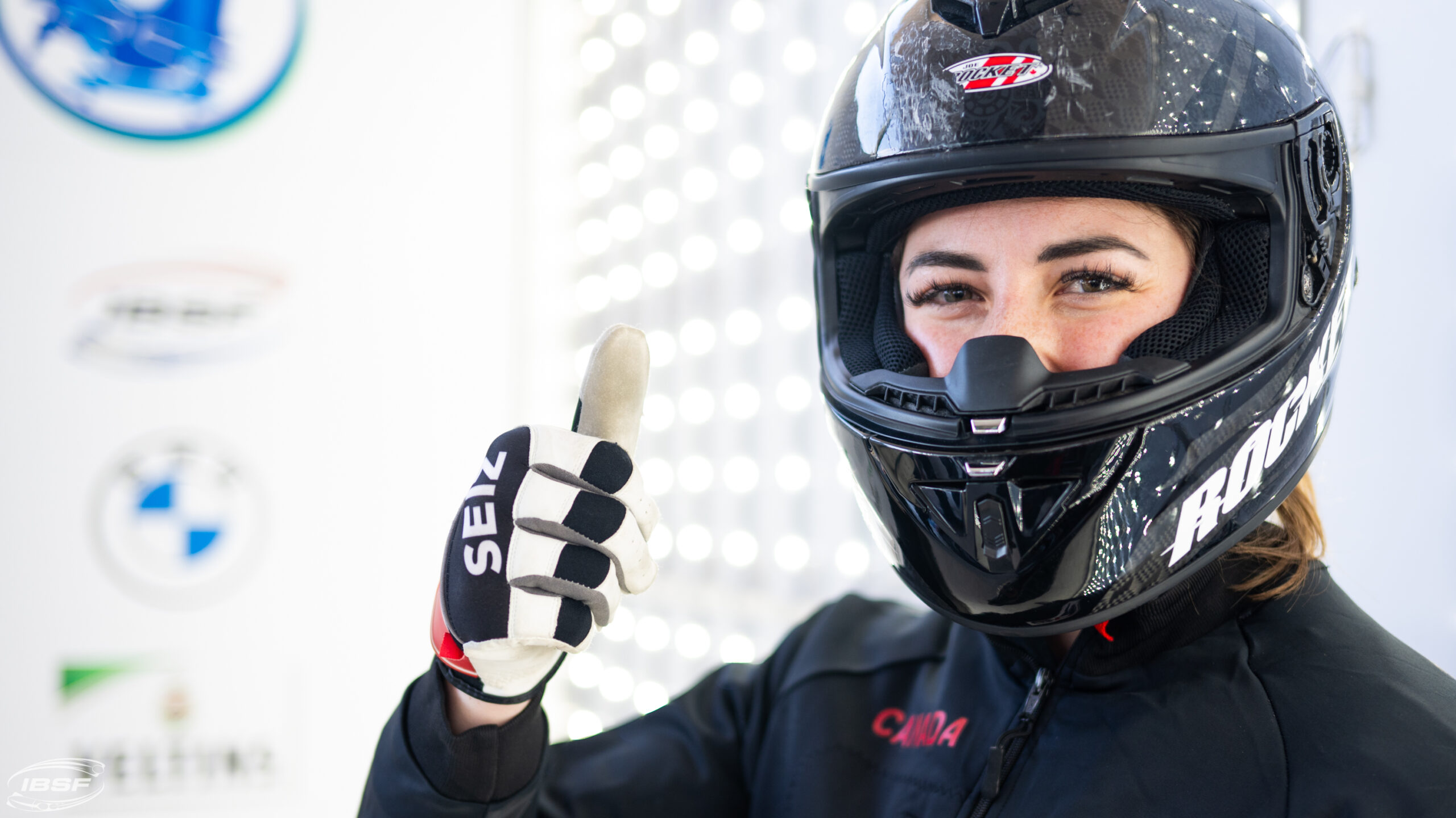 Alex Klein looks at the camera and gives a thumbs-up with her right hand. She has her bobsled helmet on, obscuring most of her face. Only her eyes are visible.