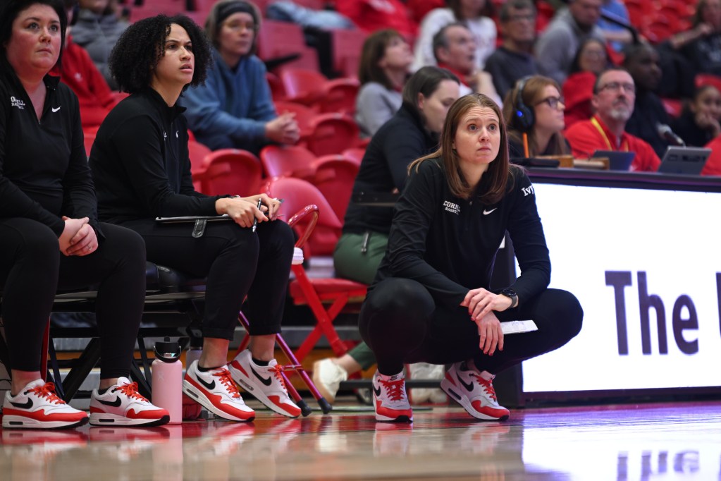 Cornell head coach Emily Garner, wearing all black, squats on the sideline in front of her team’s bench during a game.