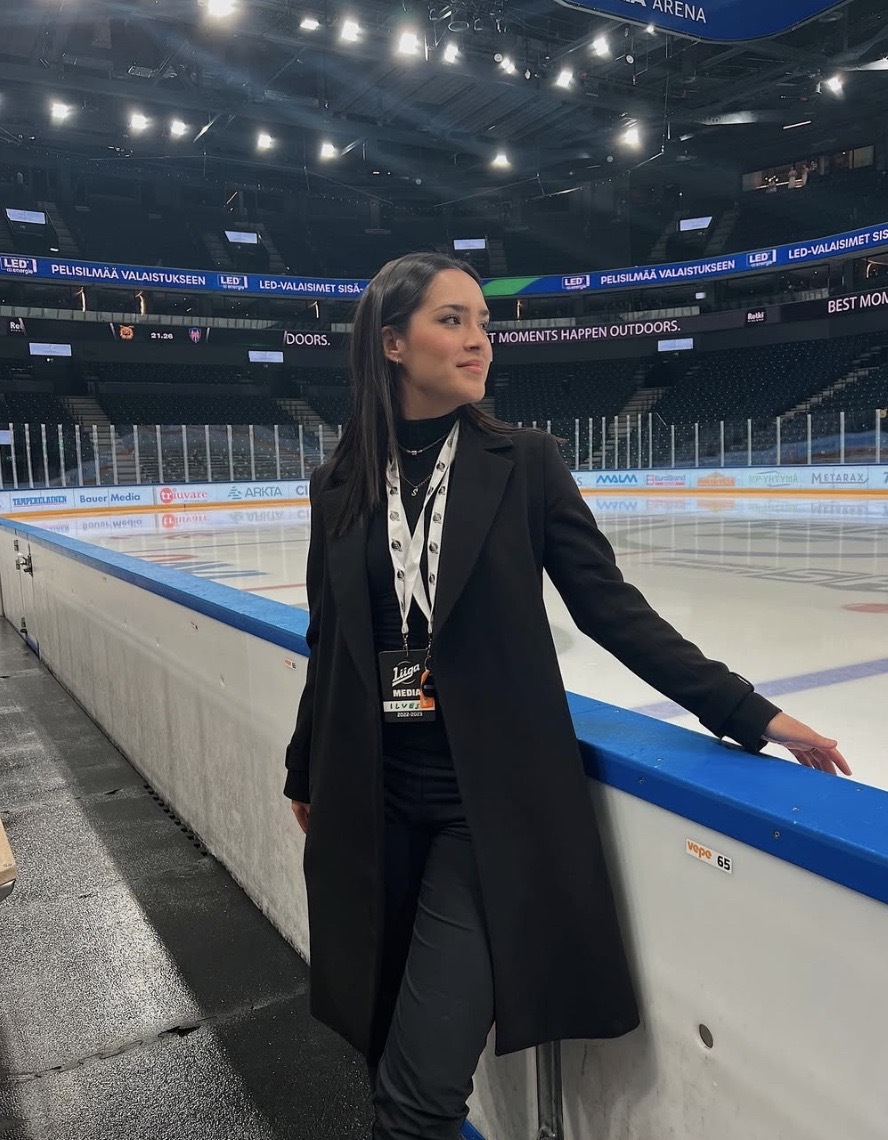 Shiayena Côté tands in front of an hockey rink looking to the right with her right arm touching the barrier.