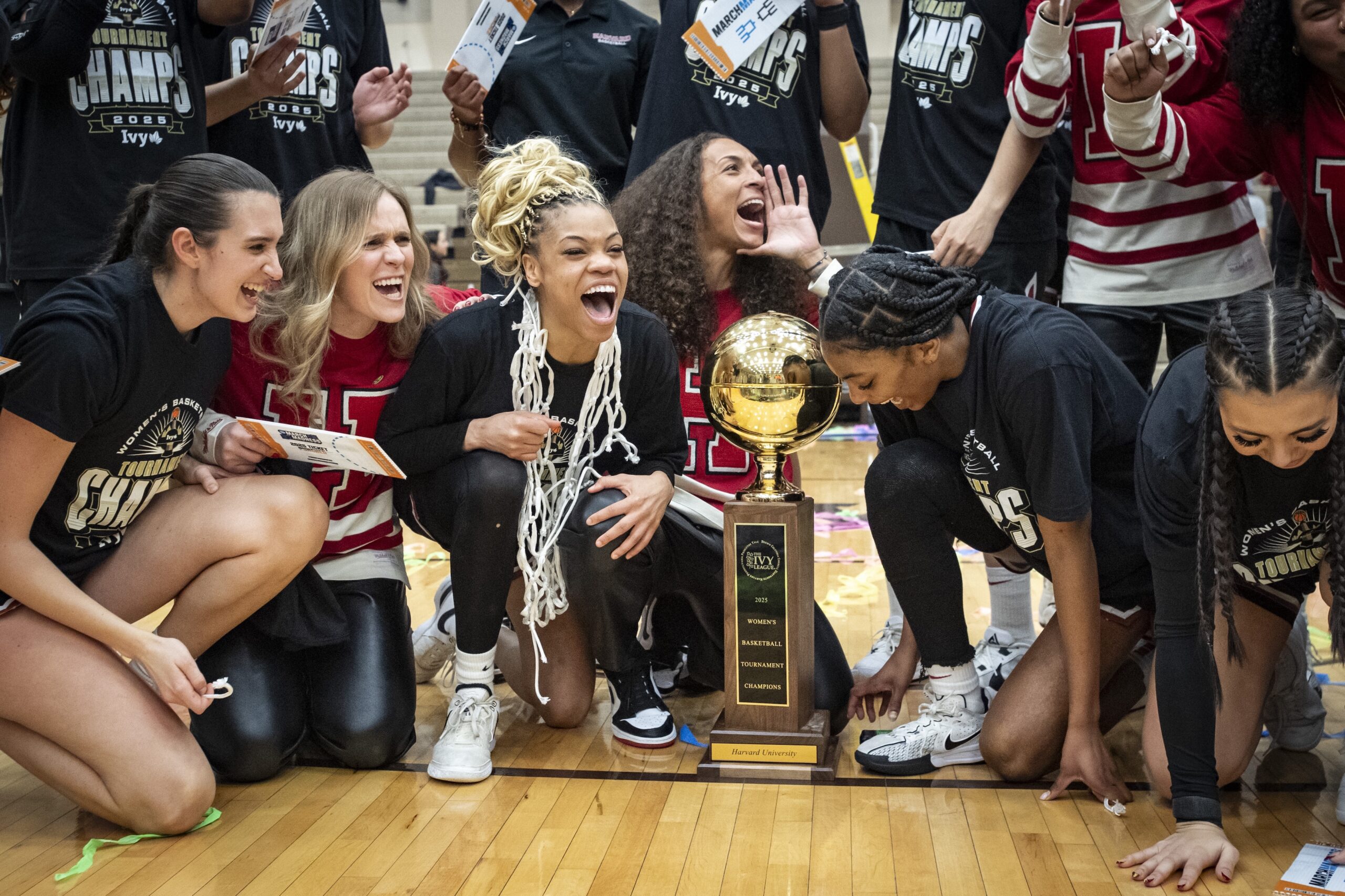Harvard players and coaches kneel on the court and pose for a photo with the Ivy League championship trophy. Guard Harmoni Turner shouts something while wearing a net around her neck. Head coach Carrie Moore puts one hand on the side of her mouth and shouts something while looking up to her left.
