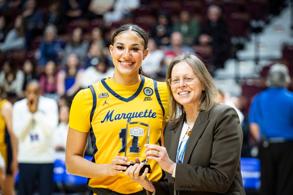 A player in a yellow Marquette jersey smiles and holds a trophy, standing next to a shorter, older woman in a blazer. 