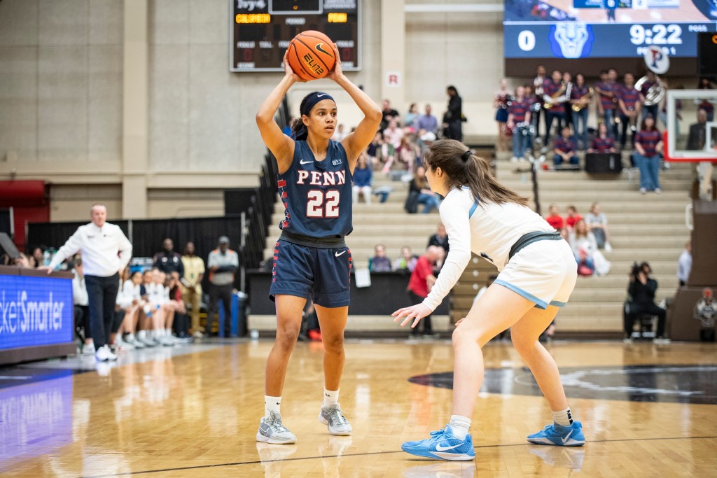 Penn guard Mataya Gayle holds the ball with two hands above her head and looks to pass from the perimeter. Columbia guard Riley Weiss is in front of her in a defensive stance with her feet on the 3-point line.