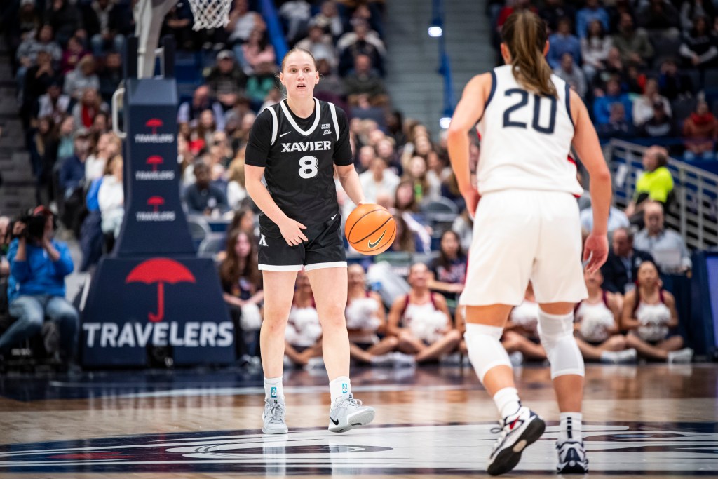 A player in a black Xavier uniform dribbles the ball while being approached by a player in a white uniform. 