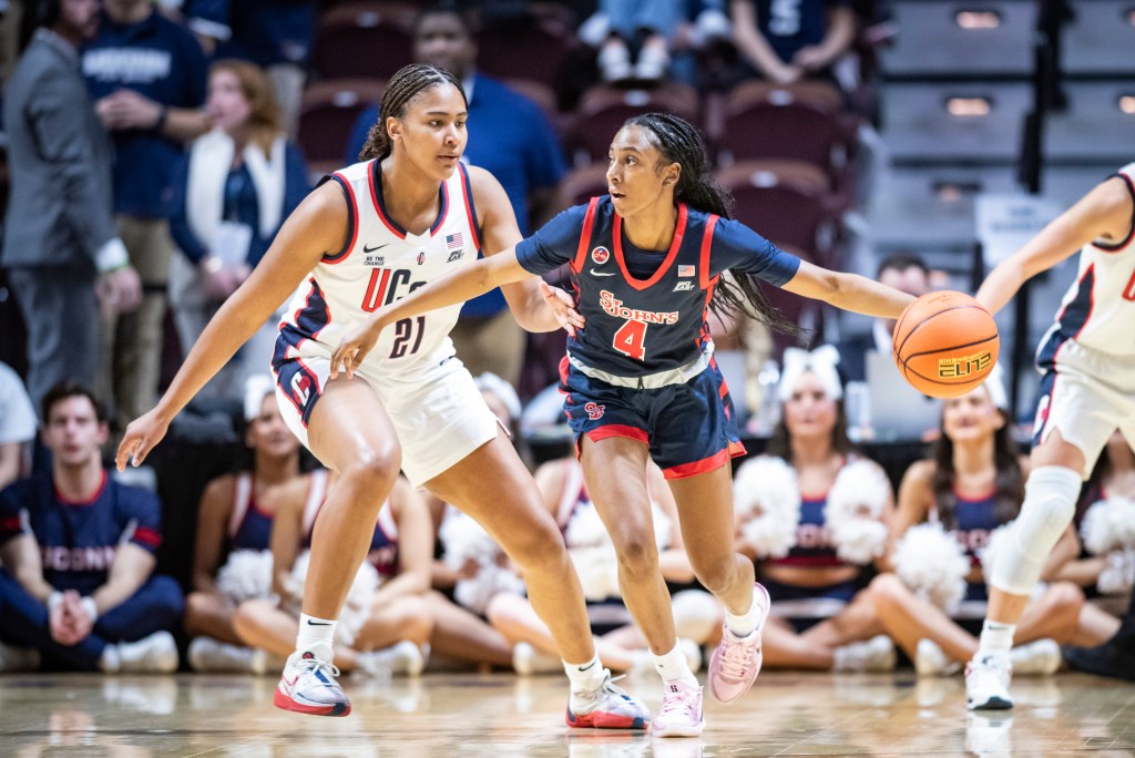 A player in a blue and red St. John's uniform dribbles the ball. To her left, a player in a white UConn uniform approaches her. The arm of a third player is also extended on the right most side of the photograph. 