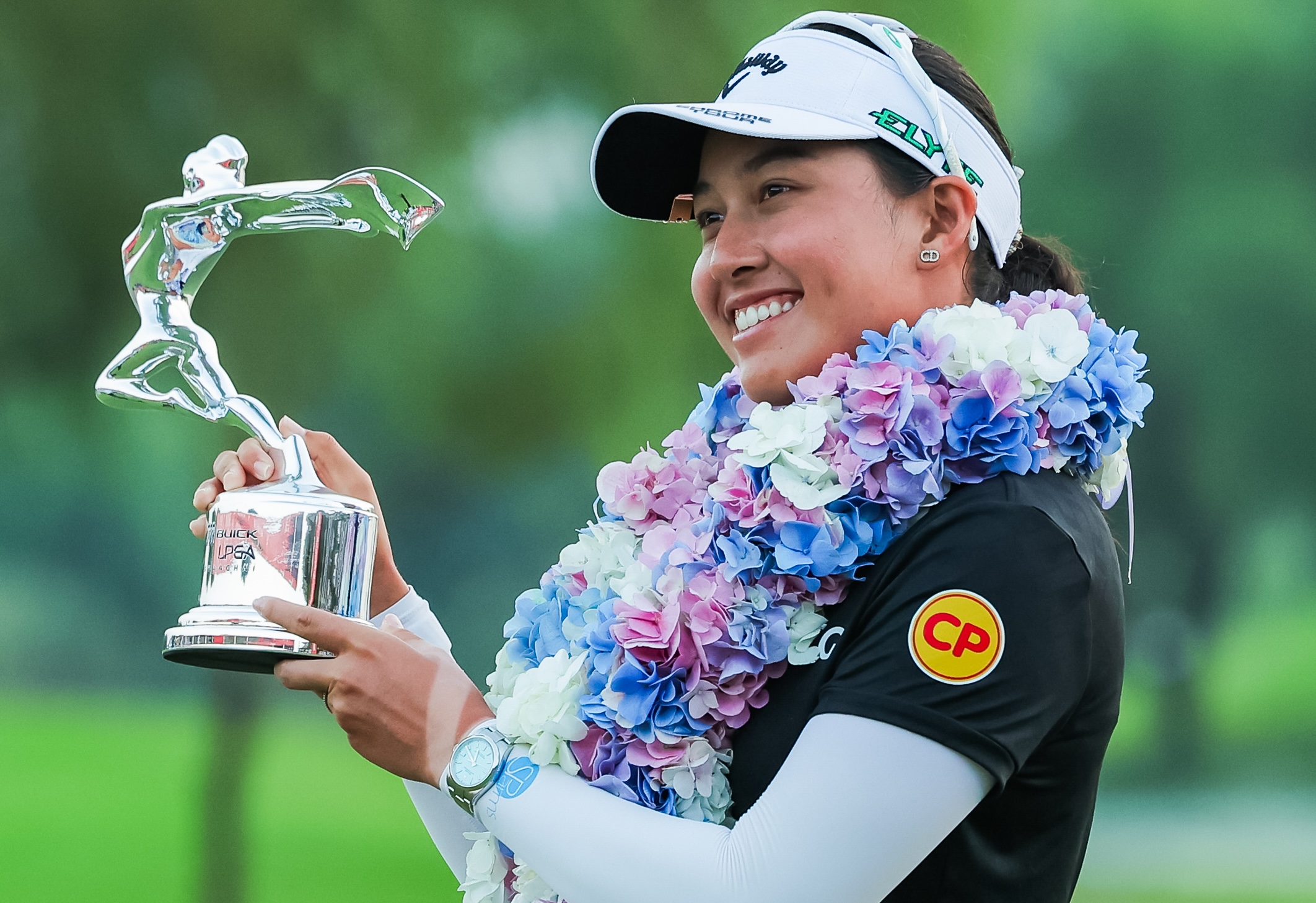 Jeeno Thitikul, winner of the 2025 Buick LPGA Shanghai, lifts the tournament trophy up to her right. She wears a large wreath of pink, purple, white, and blue flowers. She smiles and looks off at another camera.