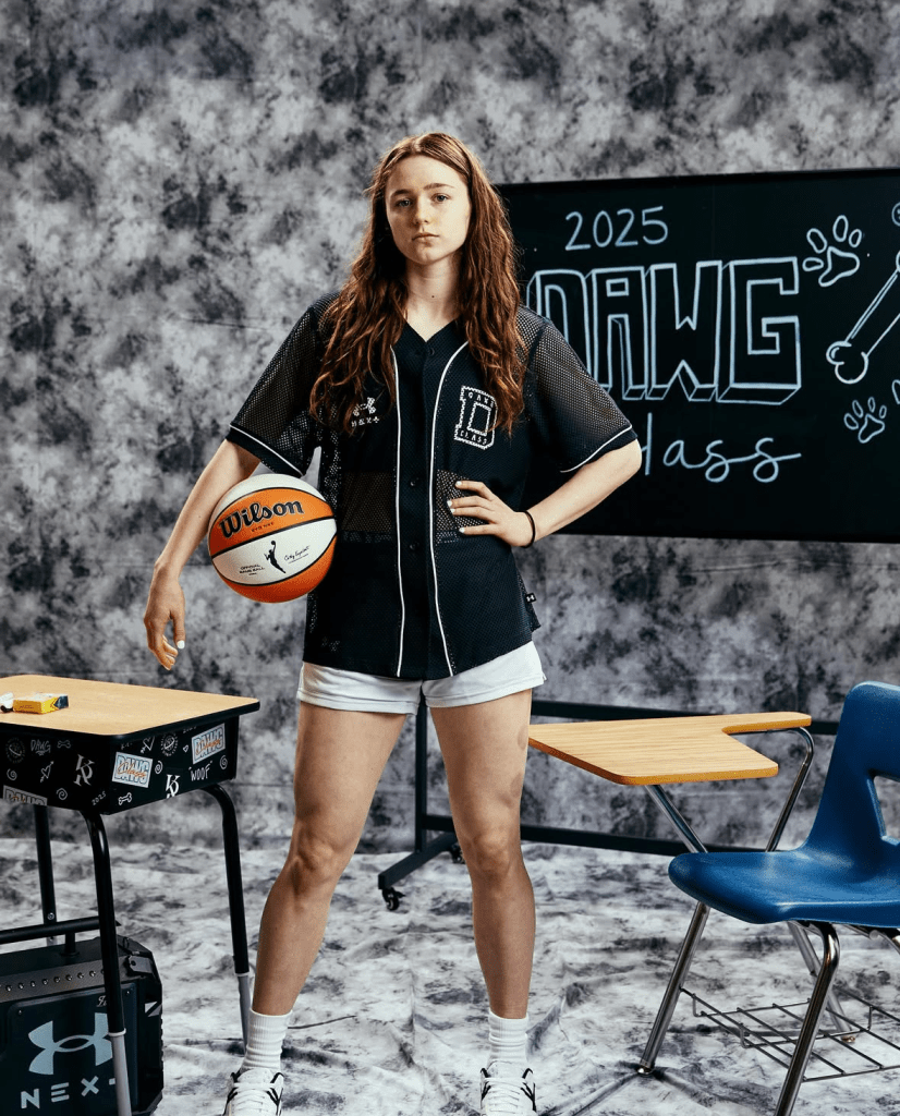 Syla Swords poses for a full body shot. She holds a basketball and wears a black jersey top and white shorts. 