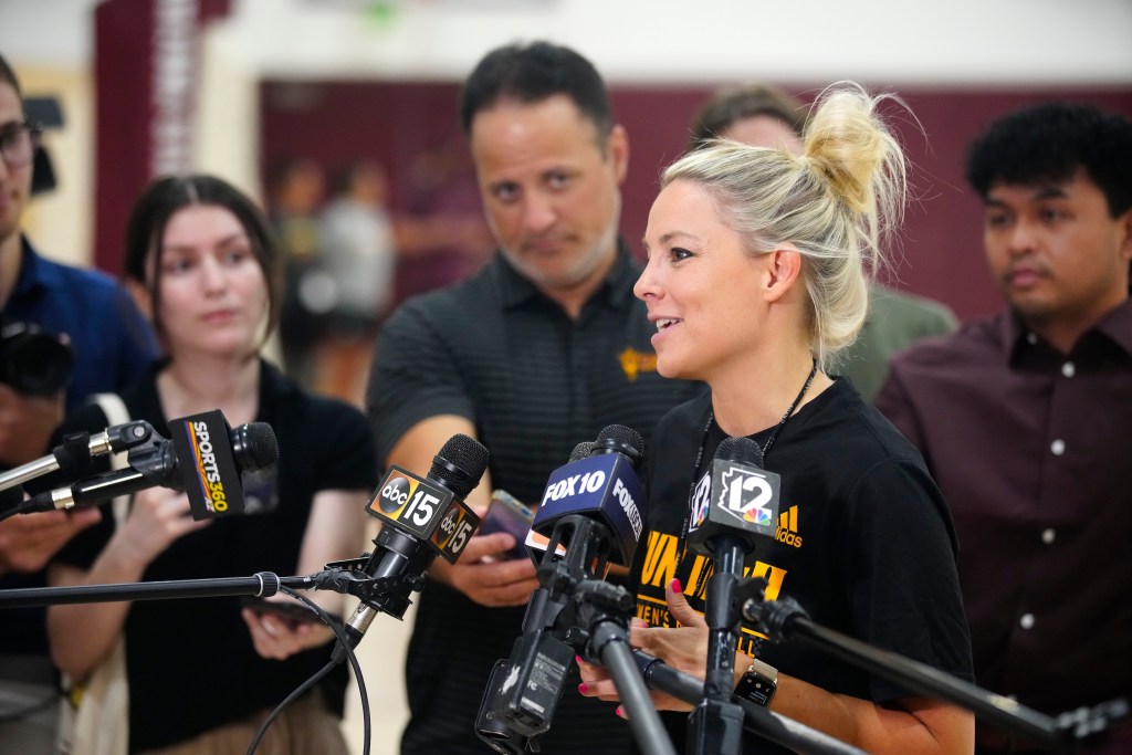 Arizona State head coach Molly Miller speaks to the press. Several microphones are positioned in front of her, and reporters surround her in the background of the photo.