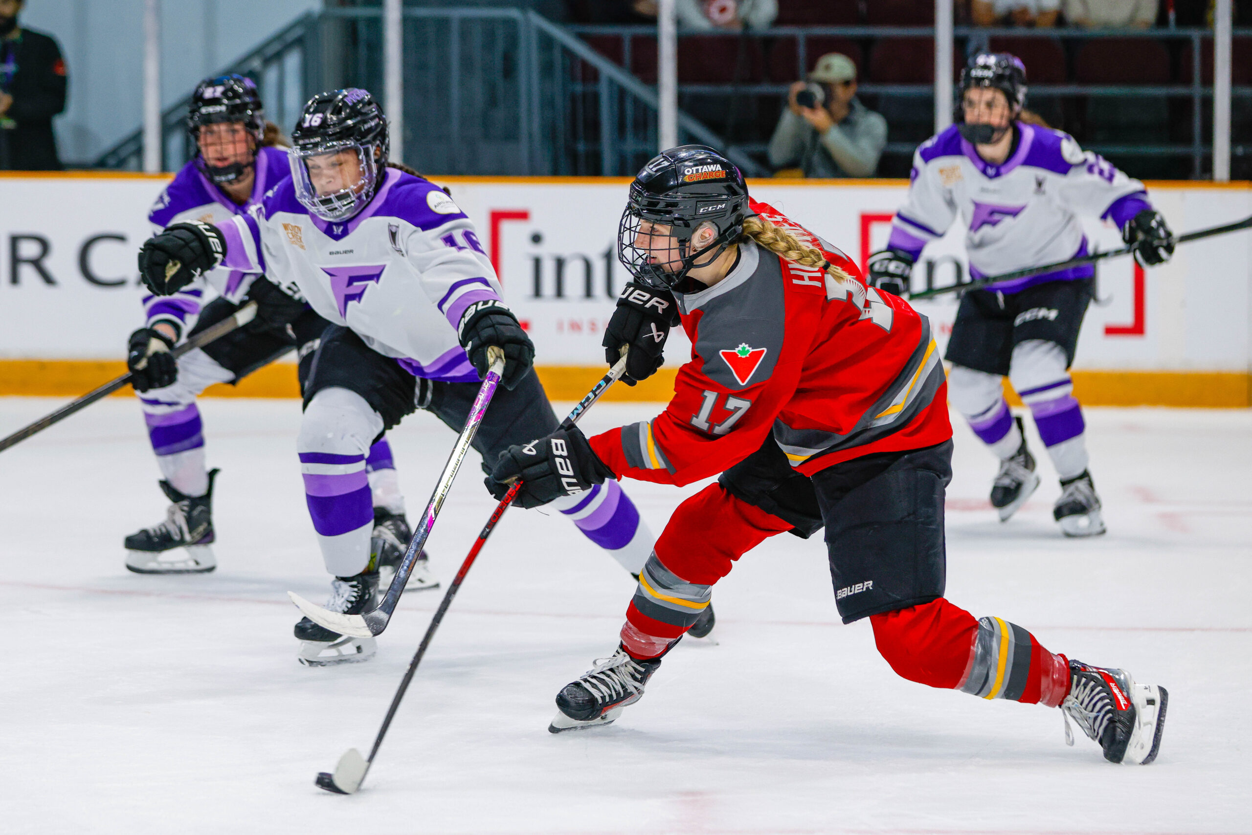 Ice hockey players skate on the ice. There are four players in the photo, two up front and two in the back. Three wear blue uniforms and one wears a red uniform.