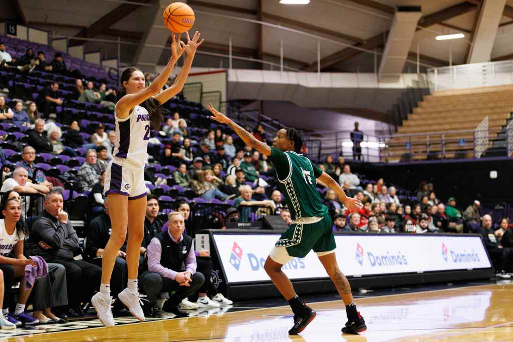Portland Forward Dyani Ananiev shoots the ball over a defender from the University of HawaiโI. She and the defender are shown from the side with an out of focus crowd behind them.