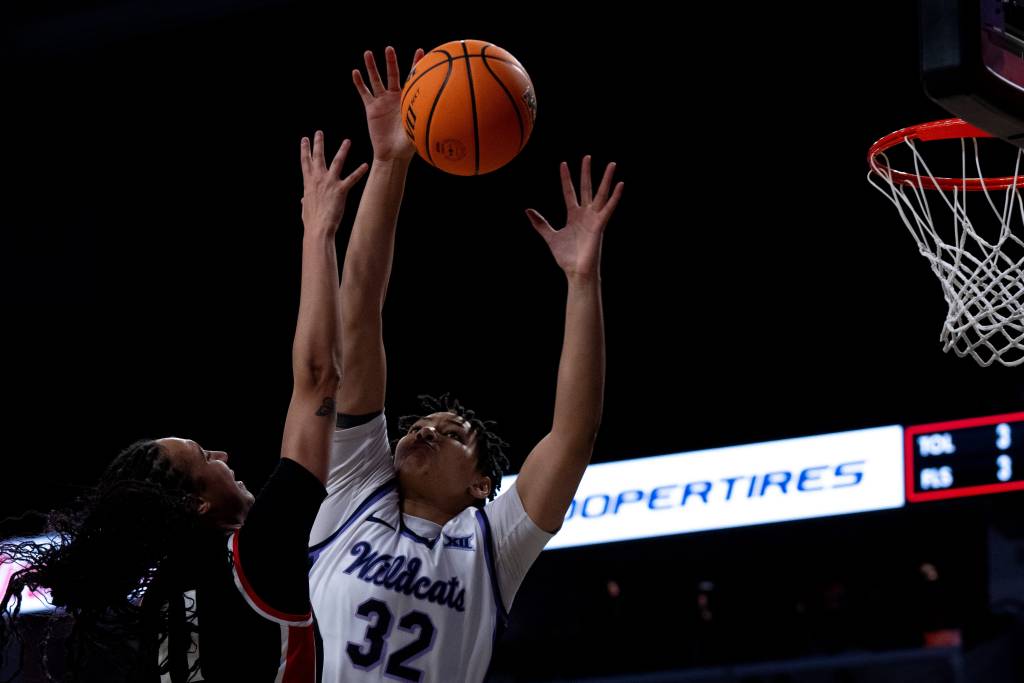 Basketball player Imani Lester blocking a shot from an opposing player. Lester wearing a white and purple basketball jersey that says "Wildcats" with the number 32. Both hands up in the air blocking a layup attempt at the basket.