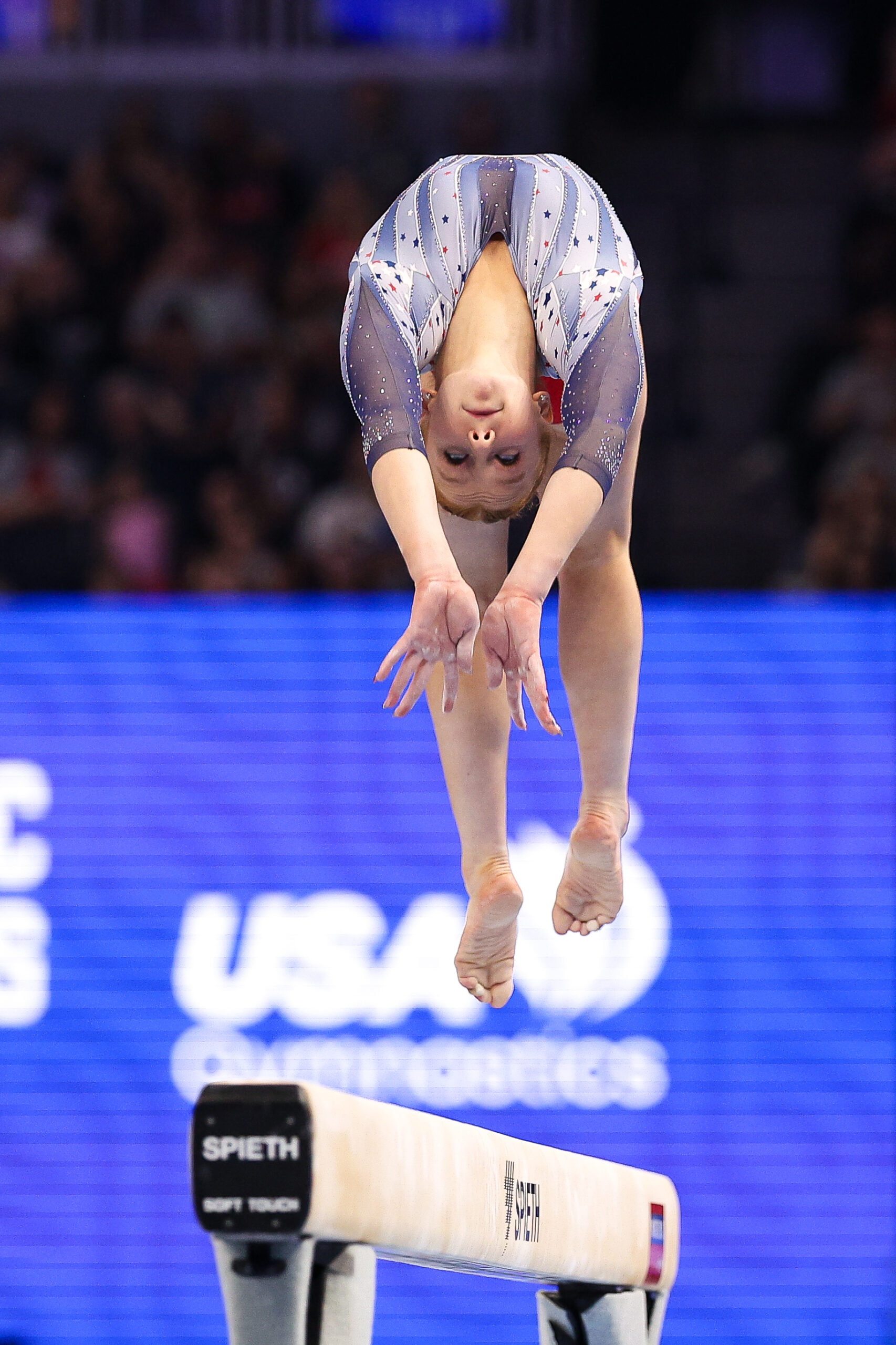 A gymnast in a red, white and blue leotard performs a back handspring on the balance beam. She is shown in midair with her back arched, moving toward the camera.