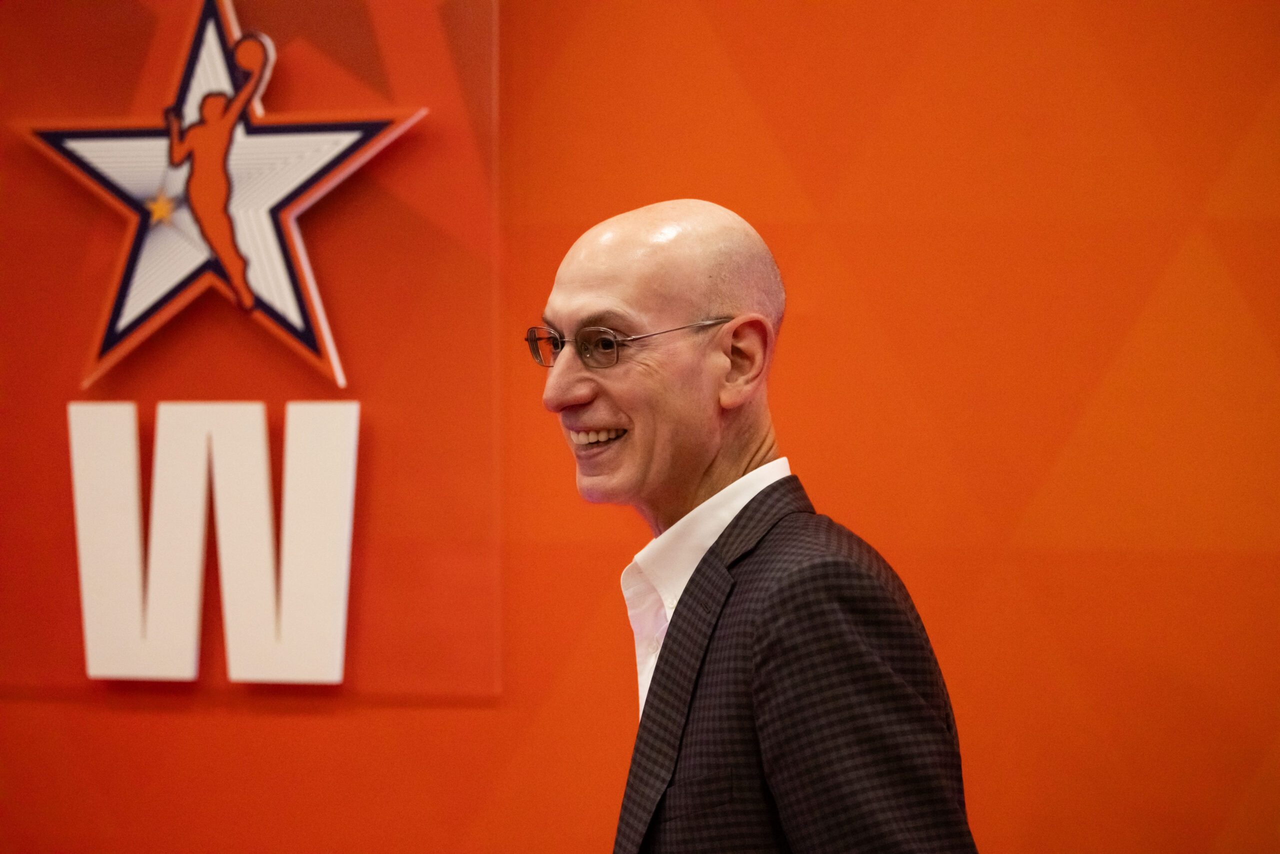 NBA commissioner Adam Silver arriving to the 2024 WNBA all-star game, walking left to right in the foreground. In the background there is an orange wall featuring the WNBA all-star logo and a block letter "W".