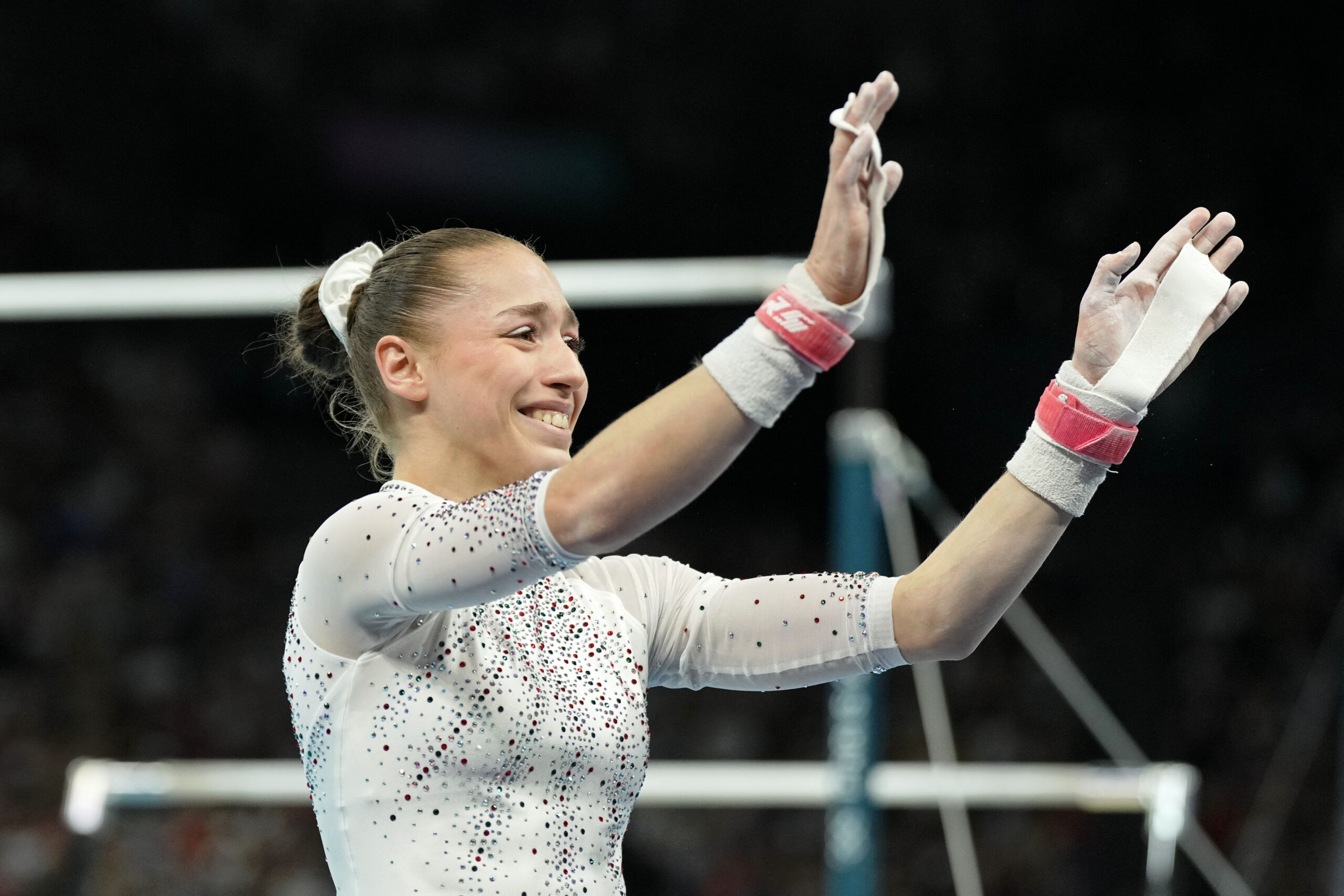 A gymnast in a white leotard waves to spectators in front of the uneven bars.