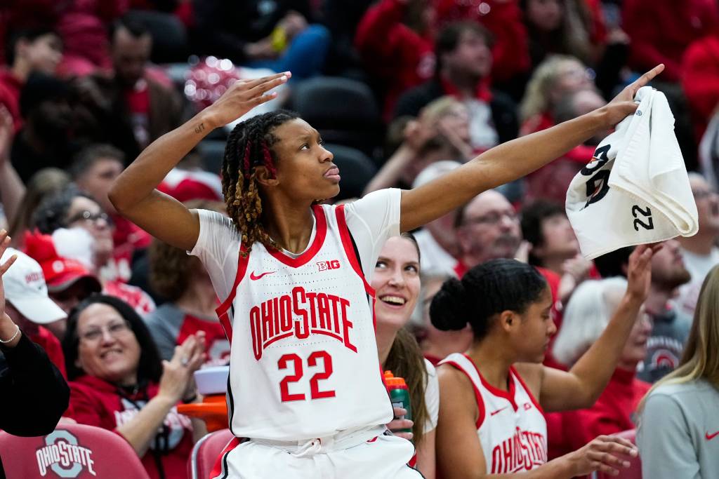 Basketball player Jaloni Cambridge wearing No. 22 in white and scarlet jersey pointing, holding a towel, as teammates and the crowd look on. Standing on the sideline. 