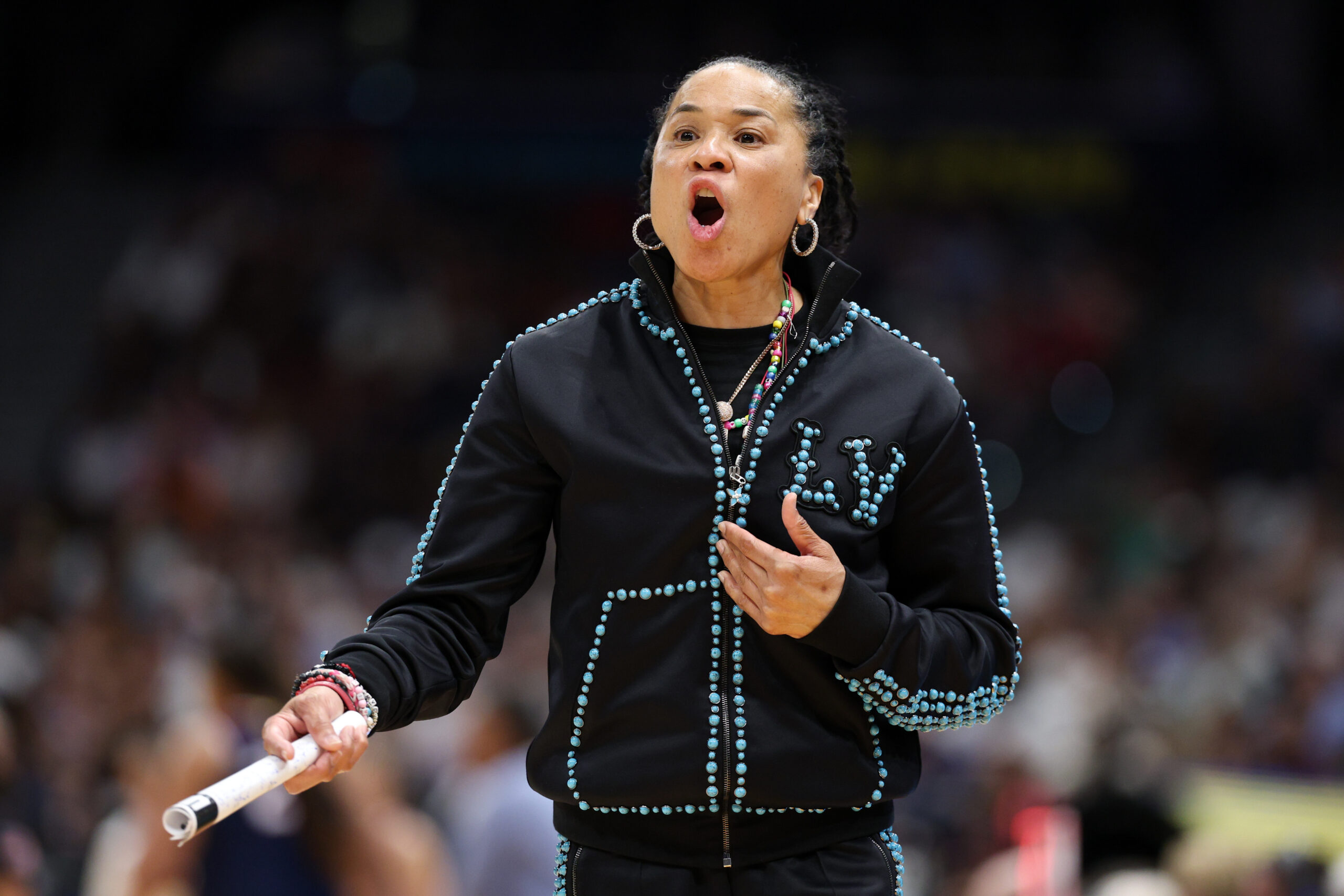 A coach shouts on the sidelines of a basketball court. She is wearing a black suit with blue trim.