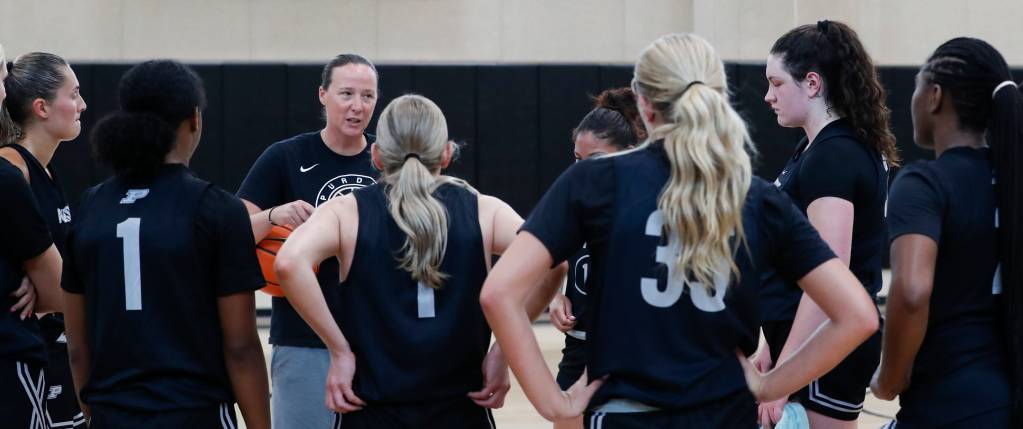 Head coach Katie Gearlds, holding a basketball under her right arm, looking at a group of eight players, with their backs facing the camera. Gearlds is talking to the players during a practice. 