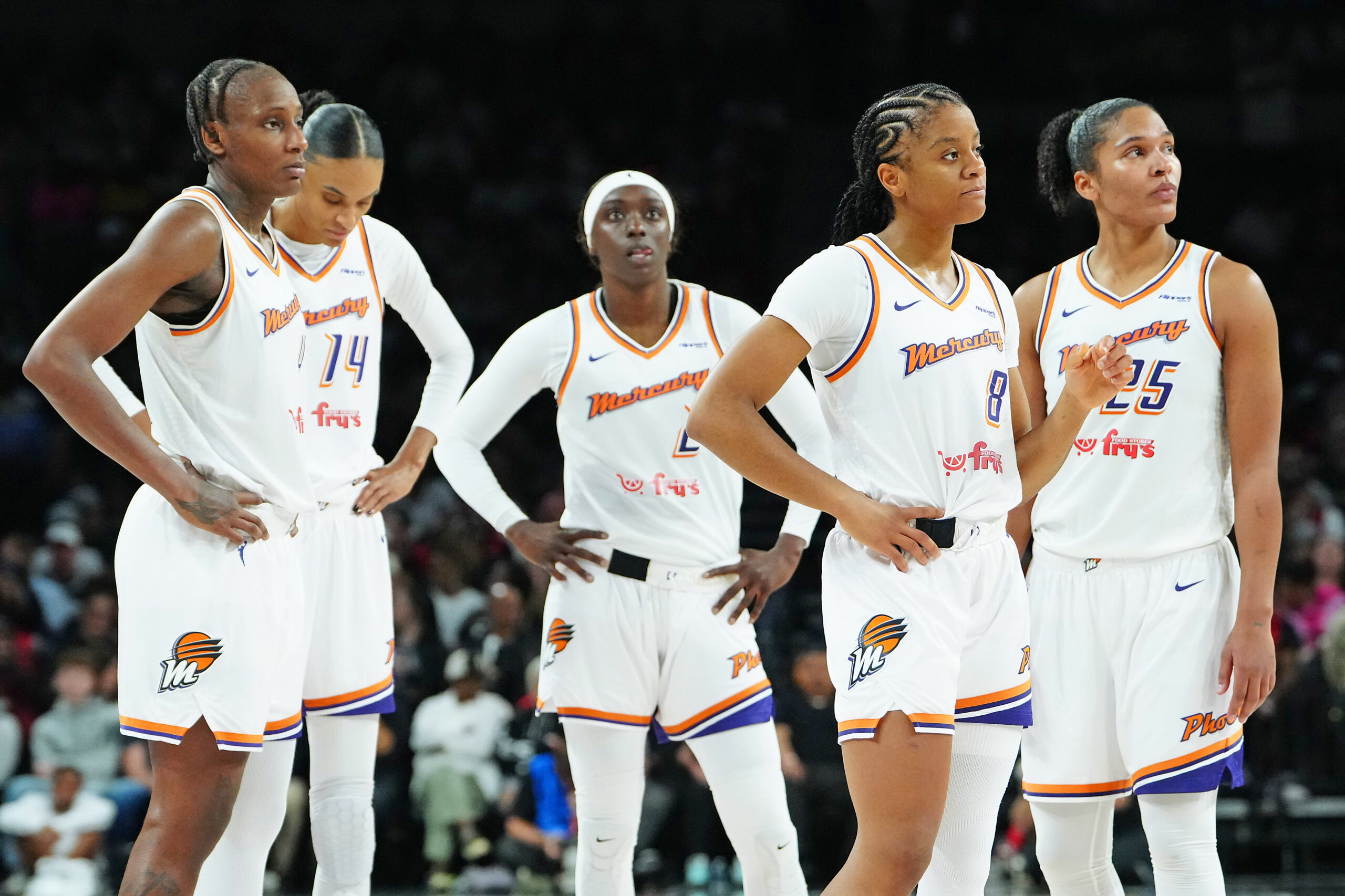 Five Phoenix Mercury players stand on the court, many of them with their hands on their hips. They are looking in different directions, and all have neutral or somber expressions on their faces.