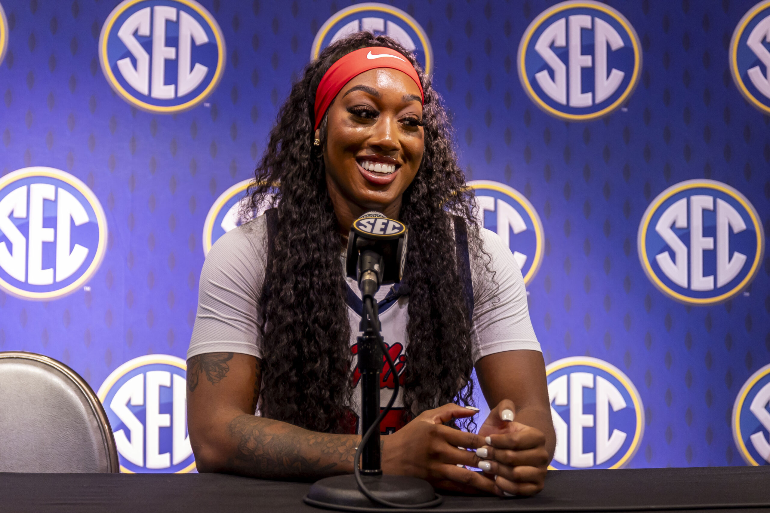 A player sits at a microphone during a press conference. She is smiling, wearing a red headband and white shirt, with a purple SEC backdrop behind her.