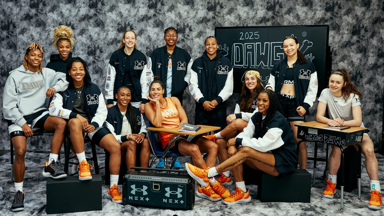 12 Basketball players sitting in front of a white and gray backdrop, posing for a photo. Three sitting at desks with a stereo sitting in front of the group that features the Under Armour "UA" logo. Blackboard in the back that says 2025 Dawg Class.