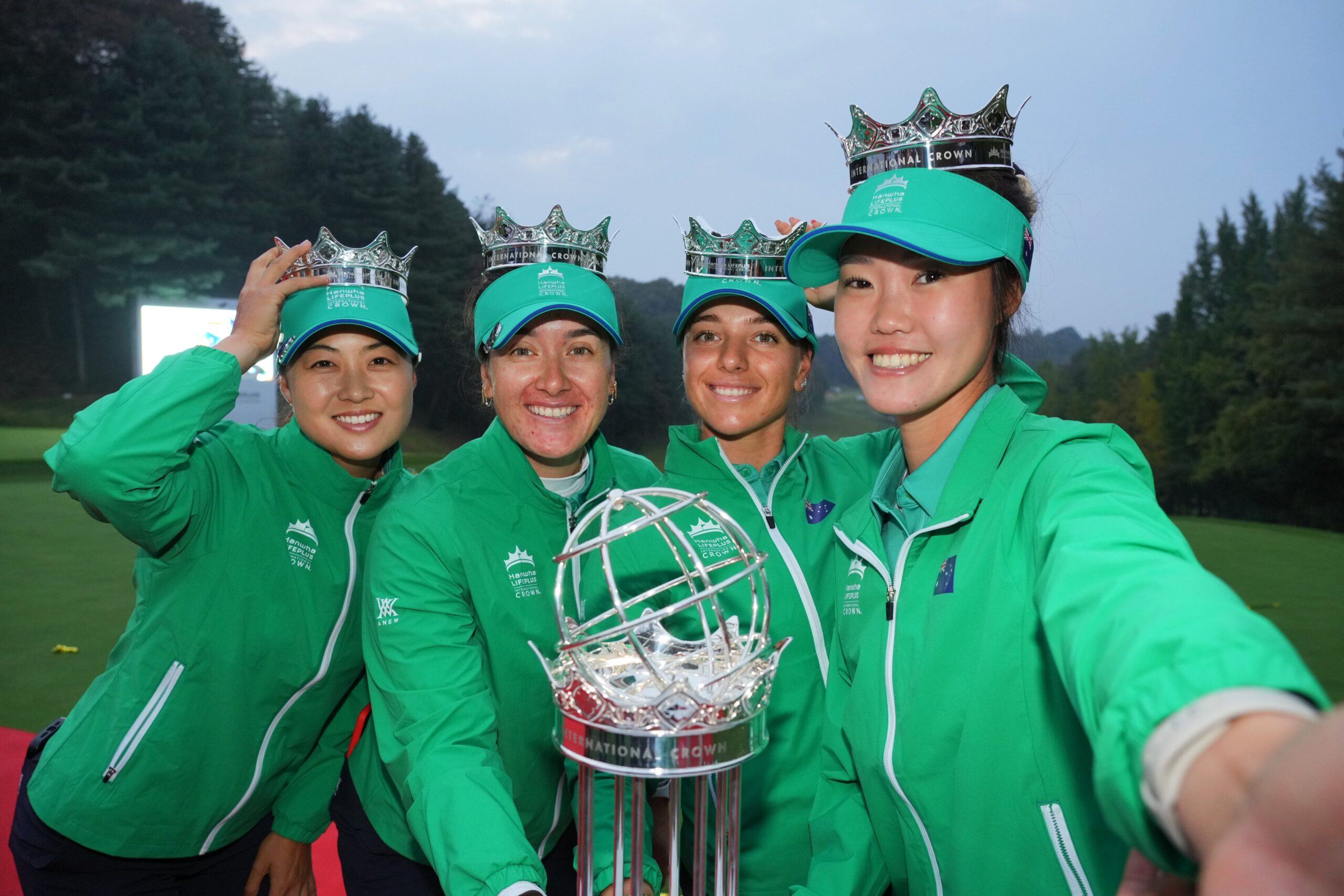 Minjee Lee, Hannah Green, Steph Kyriacou, and Grace Kim (left to right) from Team Australia pose for a selfie with the International Crown sitting in the middle of the group. They wear green golf outfits for their country and silver crowns on their heads. Lee holds hers to the top of her head. Kim's arm is outstretched taking the selfie. They all smile.