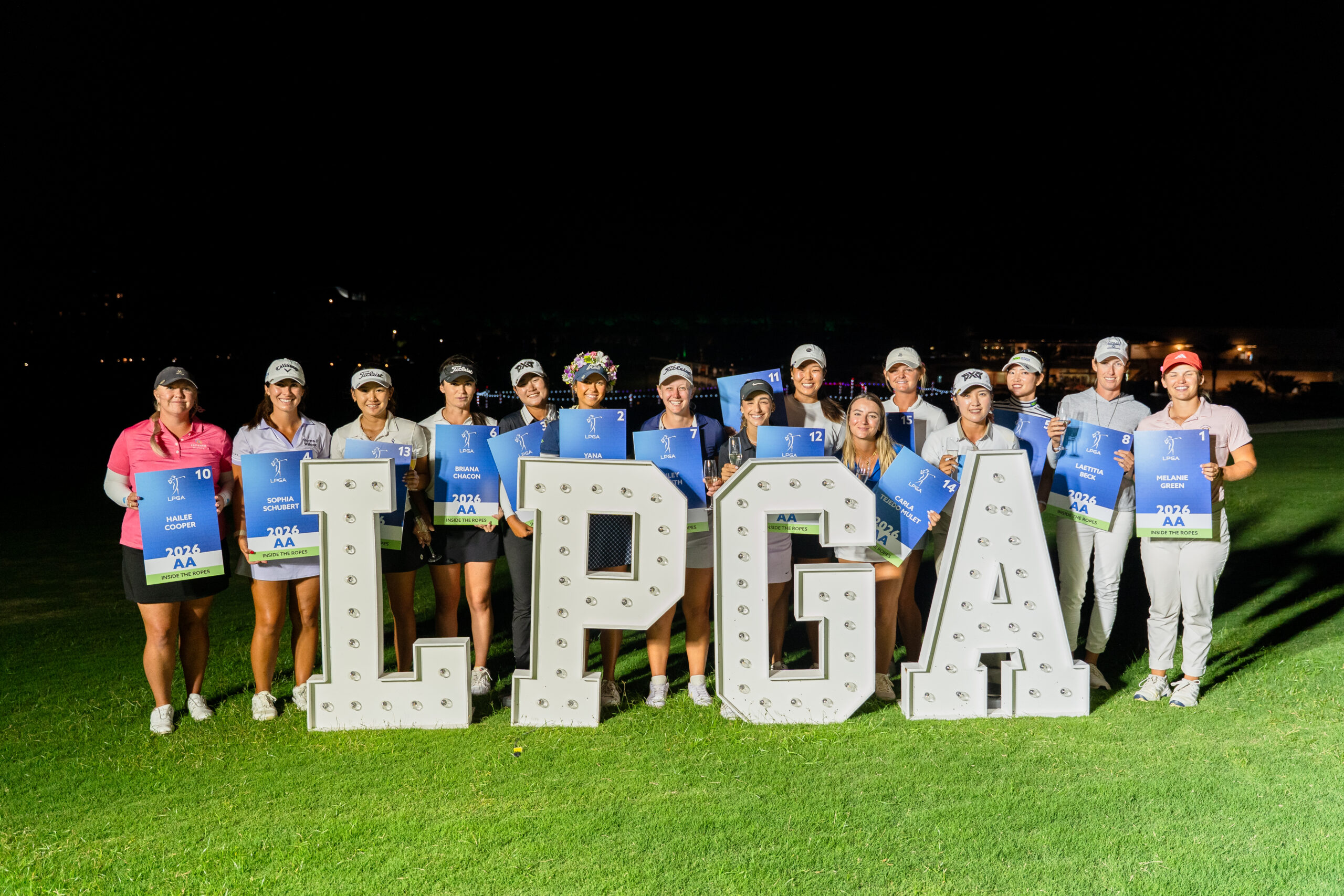Fifteen golfers stand in a row behind a giant block letter sign spelling "LPGA." They each smile and hold up signs with their names on them to signify their LPGA Tour 2026 membership.