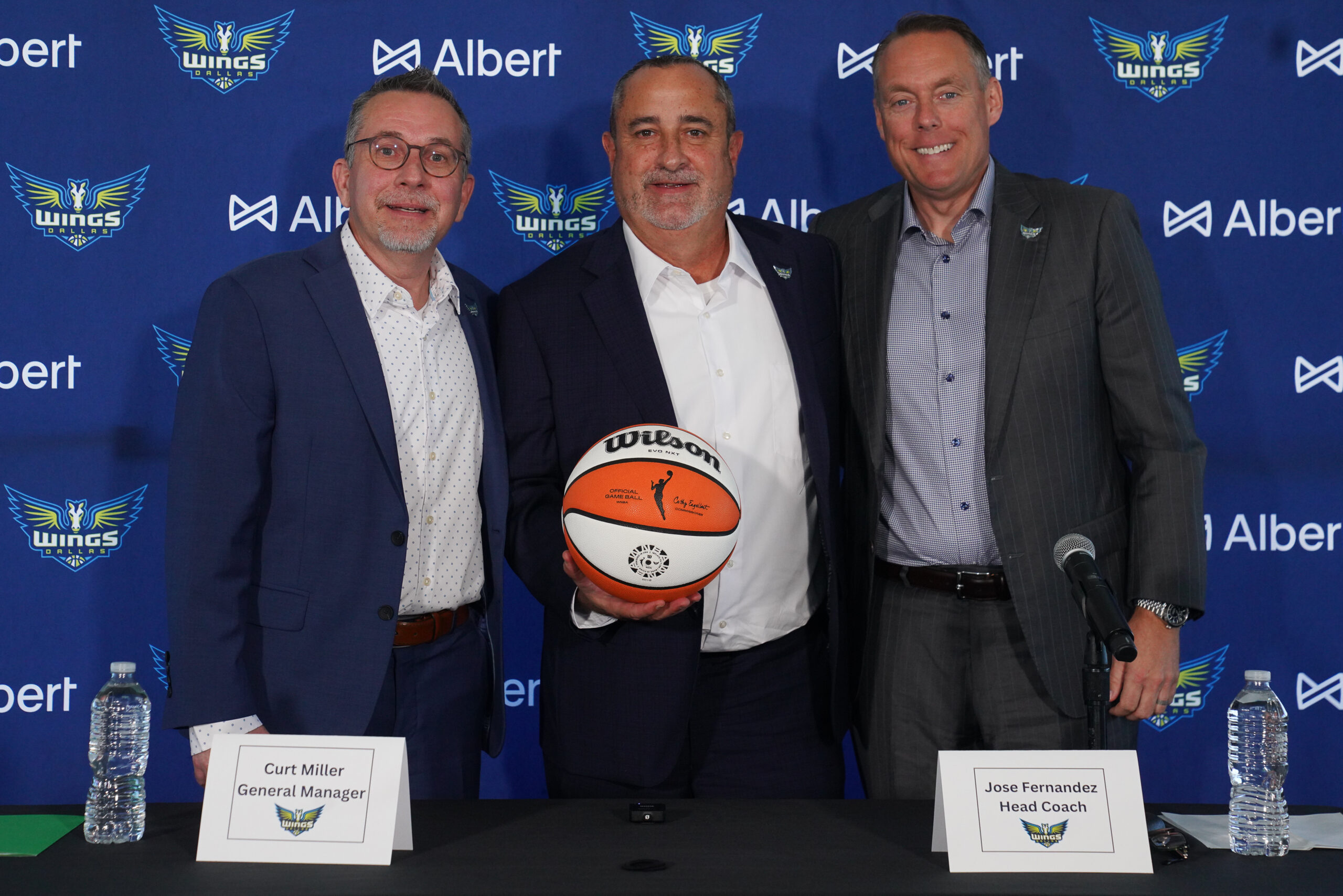 Curt Miller (left), Jose Fernandez (middle) and another man stand in front of a Dallas Wings back drop while Fernandez holds a WNBA basketball