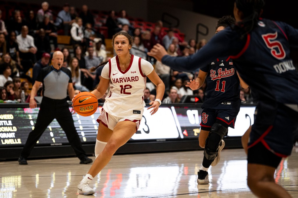 Harvard guard Karlee White has her eyes up as she dribbles the ball with her right hand just outside the lane. Her defender chases her from behind, and a help defender is sliding over.