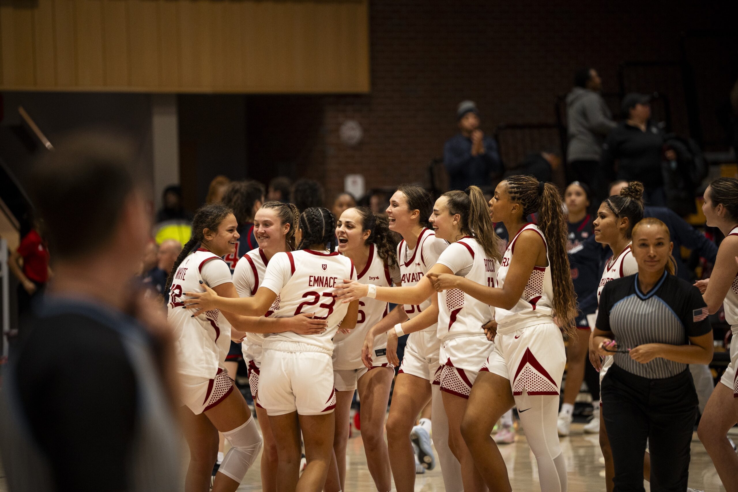 Harvard players, wearing home white uniforms, gather in a circle to celebrate a win. They are all smiling and reaching out to hug one another or pat each other on the back.