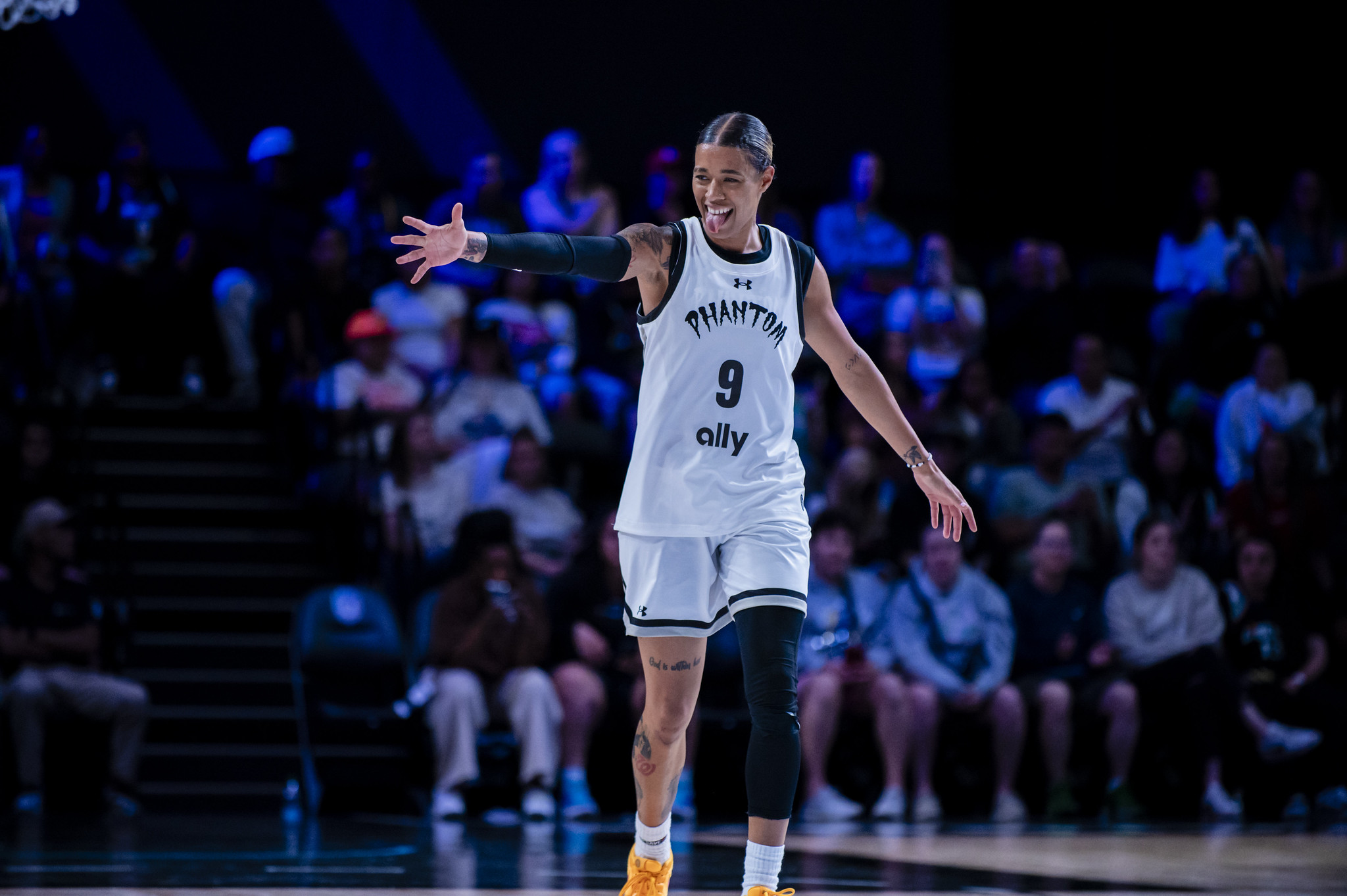 A basketball player stands on the court with her hand held out. She is wearing a white uniform and has a black protective guard on her right arm and left leg.