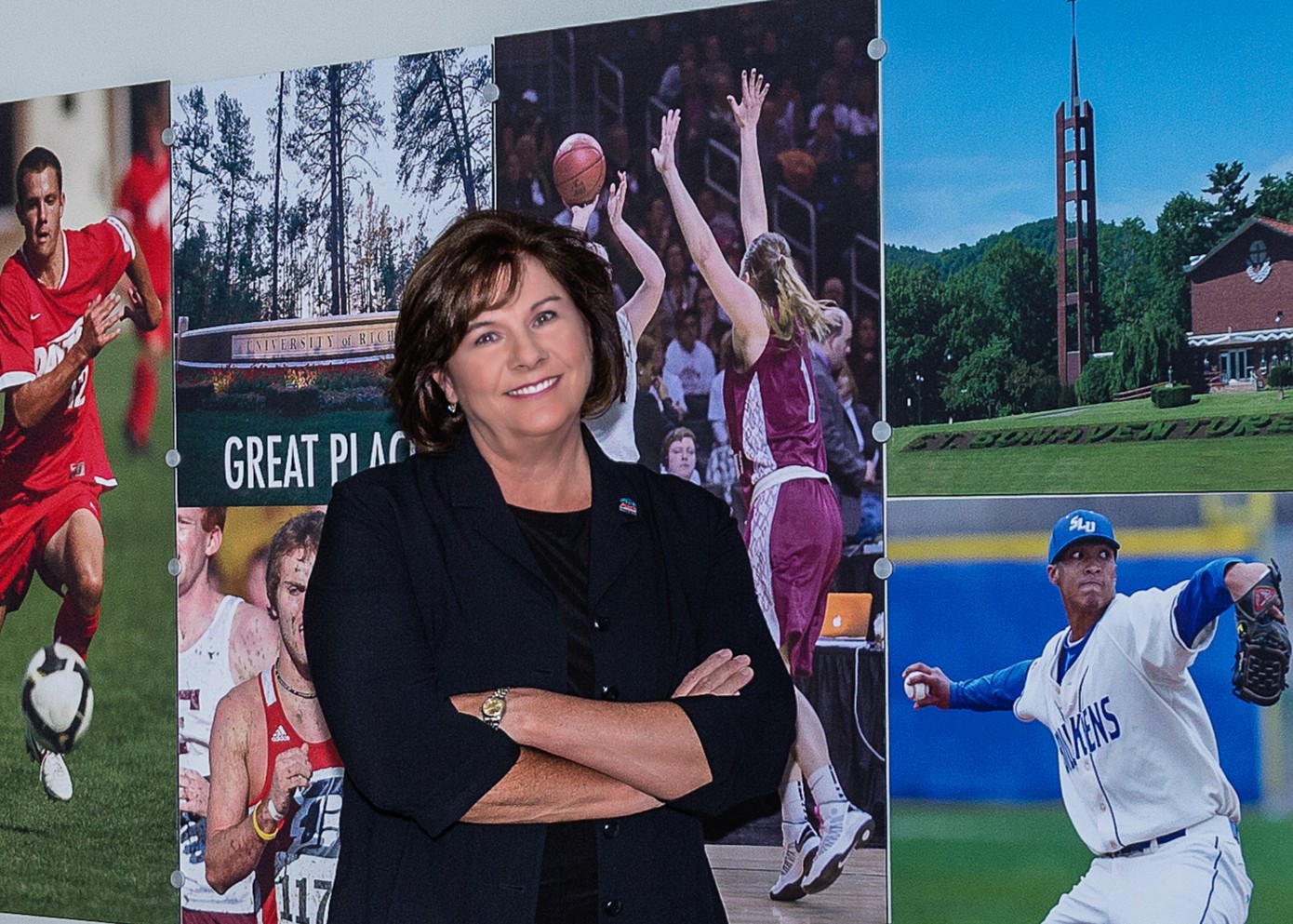 Atlantic 10 Commissioner Bernadette V. McGlade stands smiling with her arms crossed in front of a collage of photos of A-10 universities and student-athletes playing their respective sports.